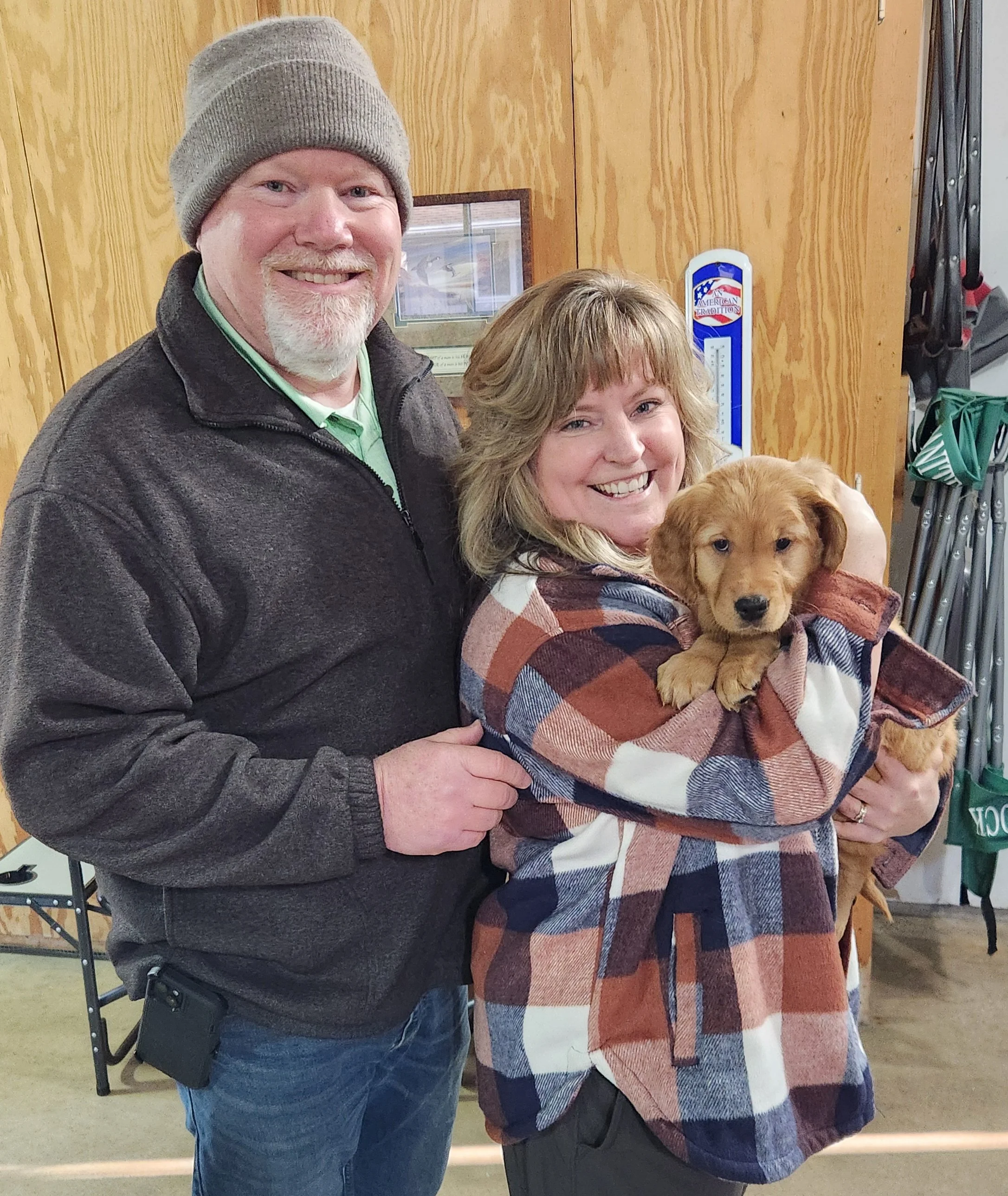 A smiling couple holding a small puppy in a plaid jacket inside a wooden room with golfing equipment on the wall.