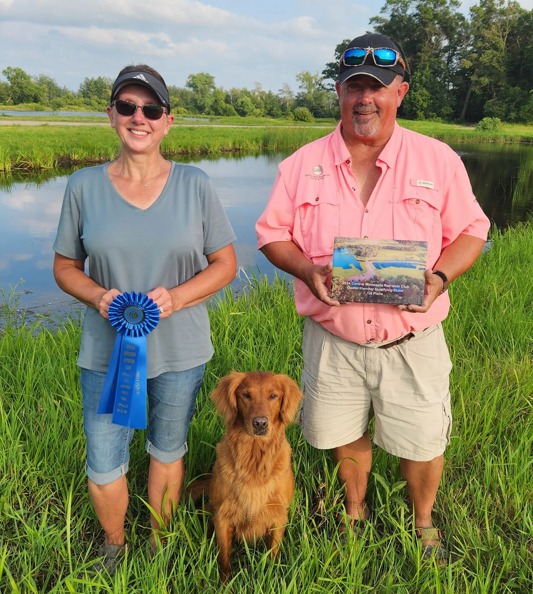 Two people and a dog standing outdoors next to a pond, with trees and grass in the background. The woman on the left is holding a blue ribbon, and the man on the right is holding a box. The dog is sitting in front of them.