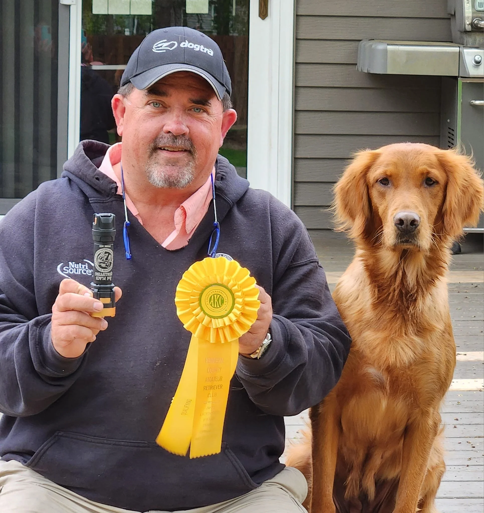 A man wearing a black Jim Dotta baseball cap and black hoodie, holding a flashlight and a yellow ribbon rosette, sitting next to a golden retriever dog on a wooden deck outside a house.