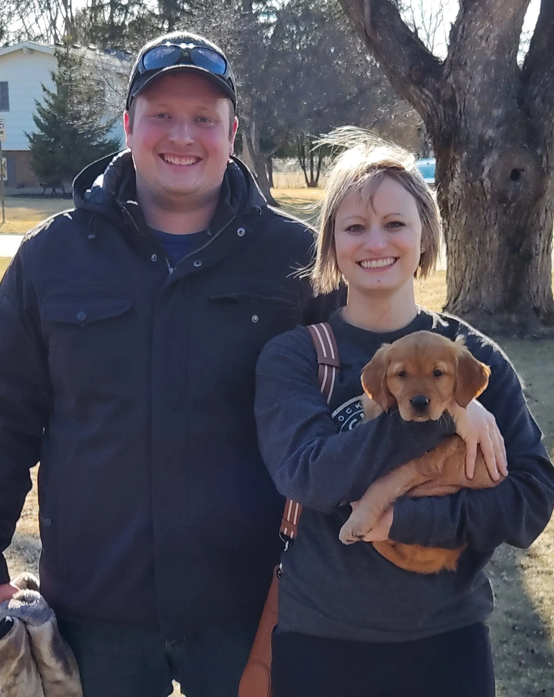 A smiling man and woman standing outdoors, with the woman holding a small brown puppy. There is a large tree and houses in the background.