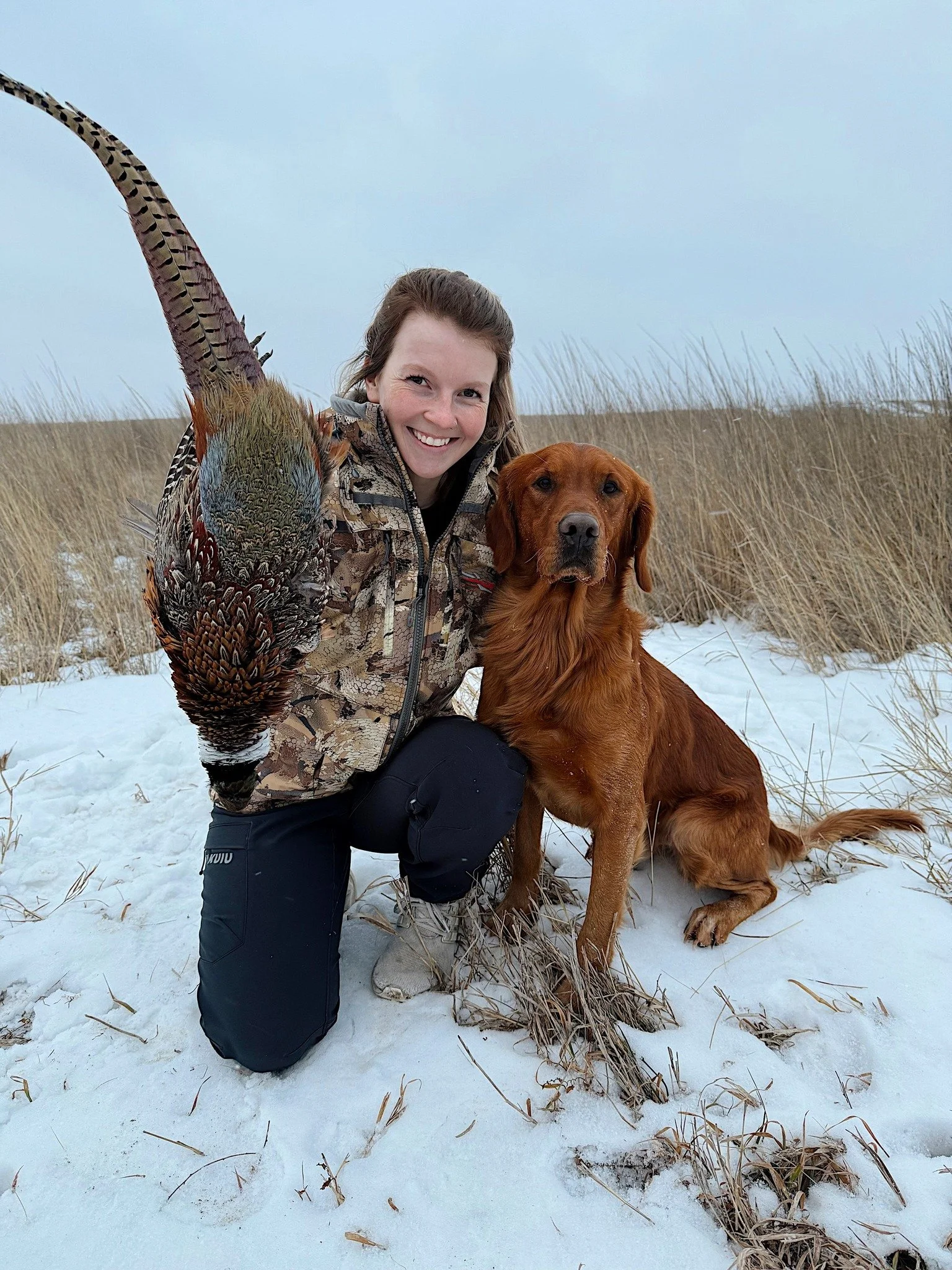 A woman holding a bird with a long tail, kneeling next to a golden retriever dog on snow-covered ground in a grassy, open landscape.