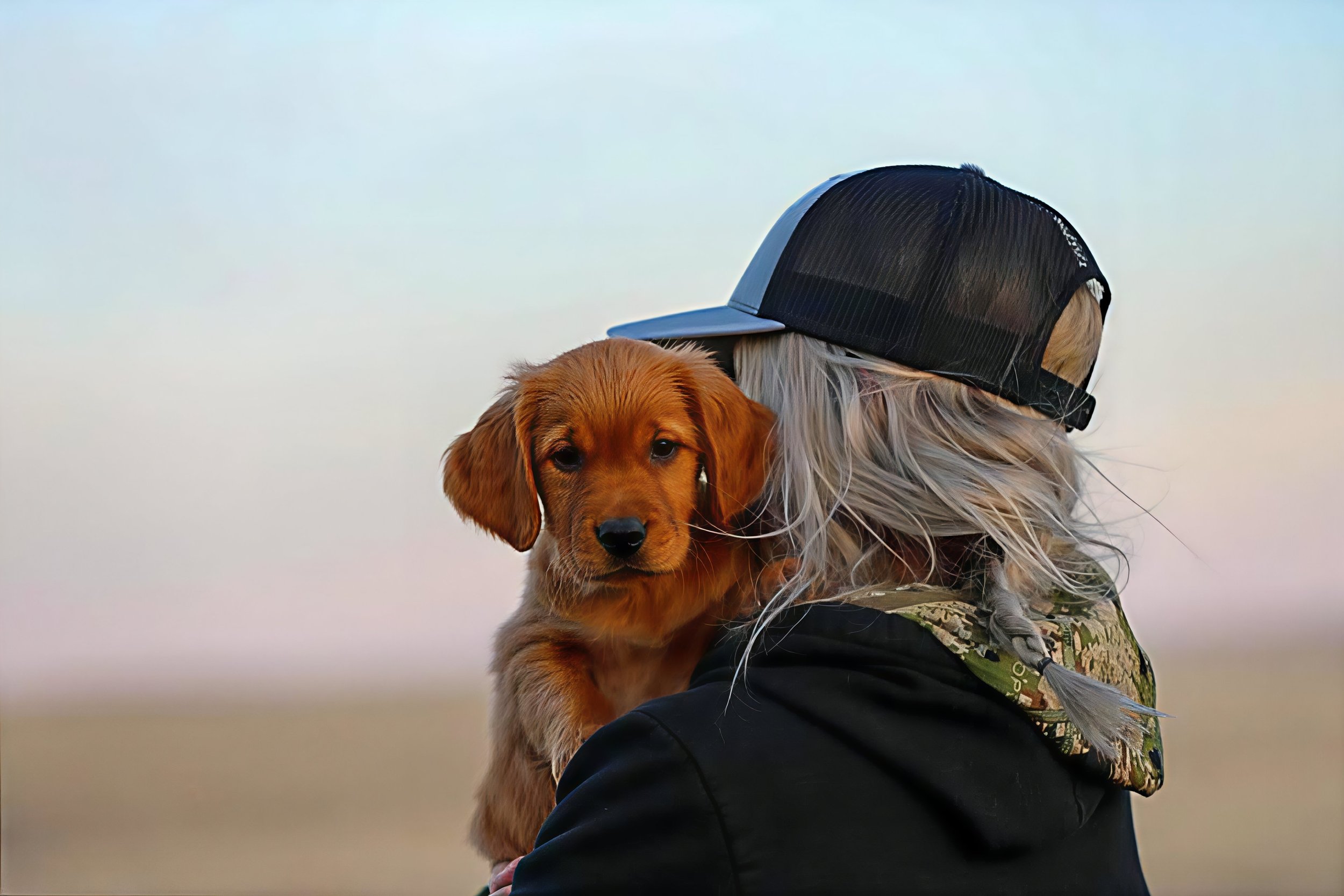 Person with long hair wearing a black and white cap holding a small golden retriever puppy outdoors during daytime.