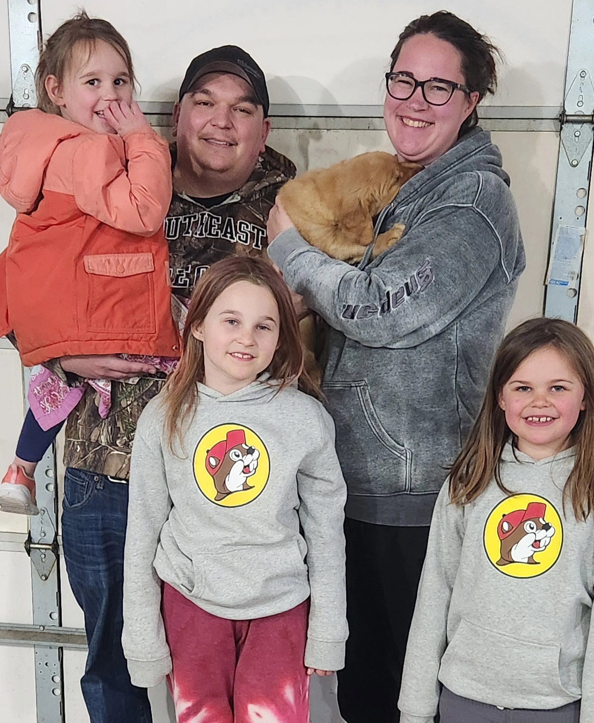 A group of five people, including two adults and three children, posing in front of a garage door, with one adult holding a puppy. All children are wearing gray hoodies with a cartoon dog logo.