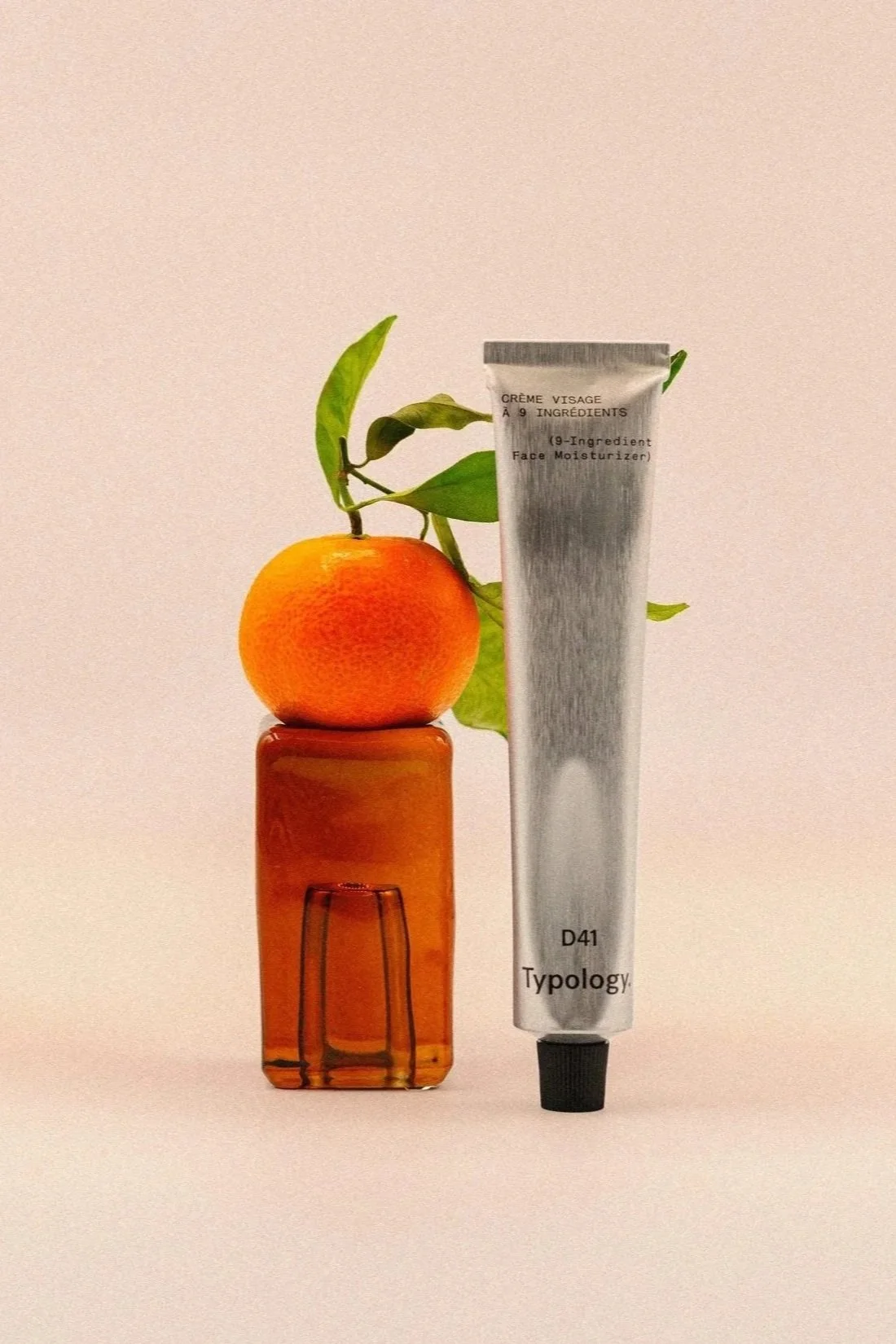 Un orange repose sur un petit vase en verre orange, à côté d'un tube de crème visage de couleur argentée. Il y a des feuilles vertes au-dessus de l'orange.