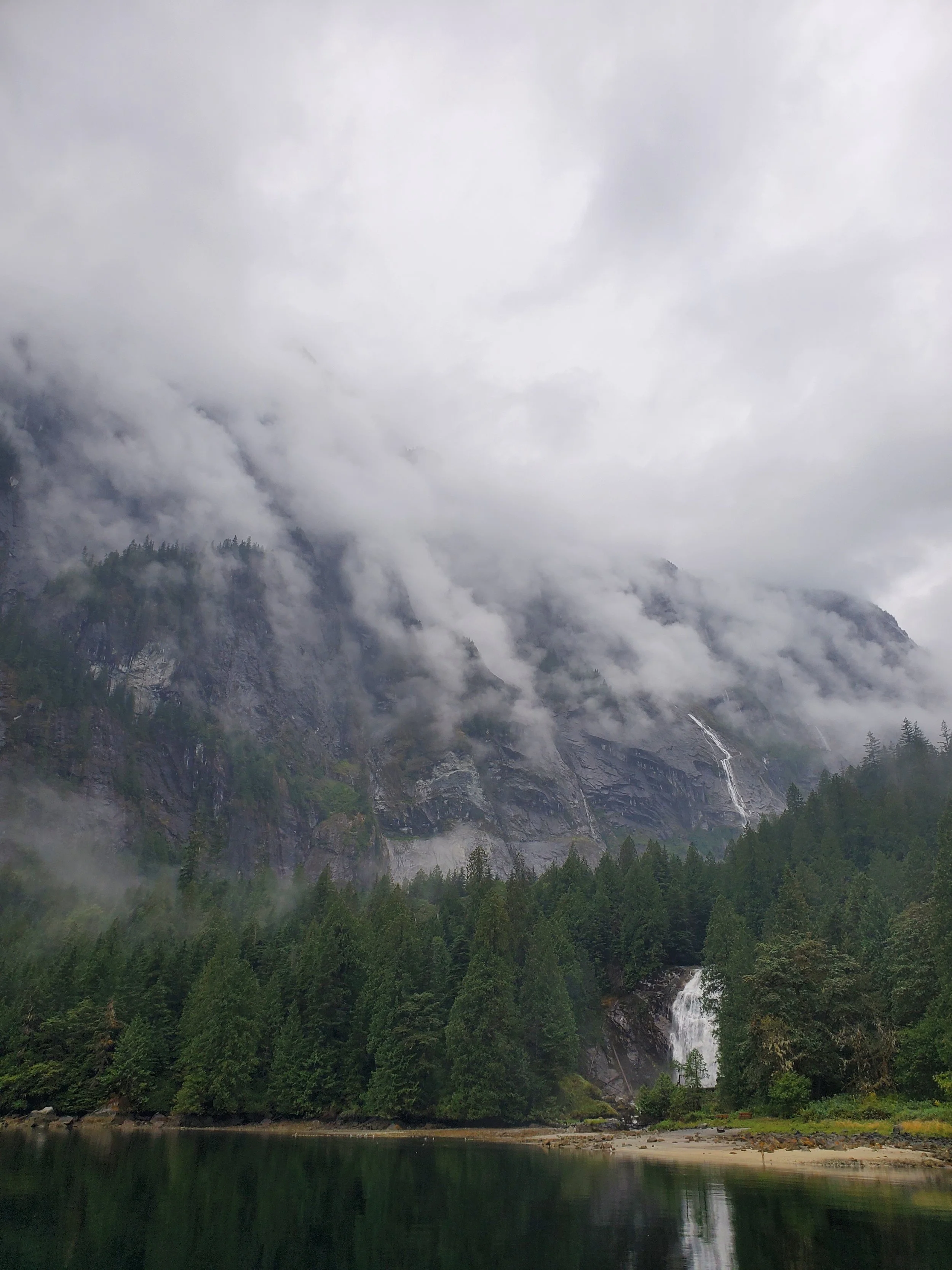Cloud-covered mountains above a forest and a waterfall flowing into a calm lake.