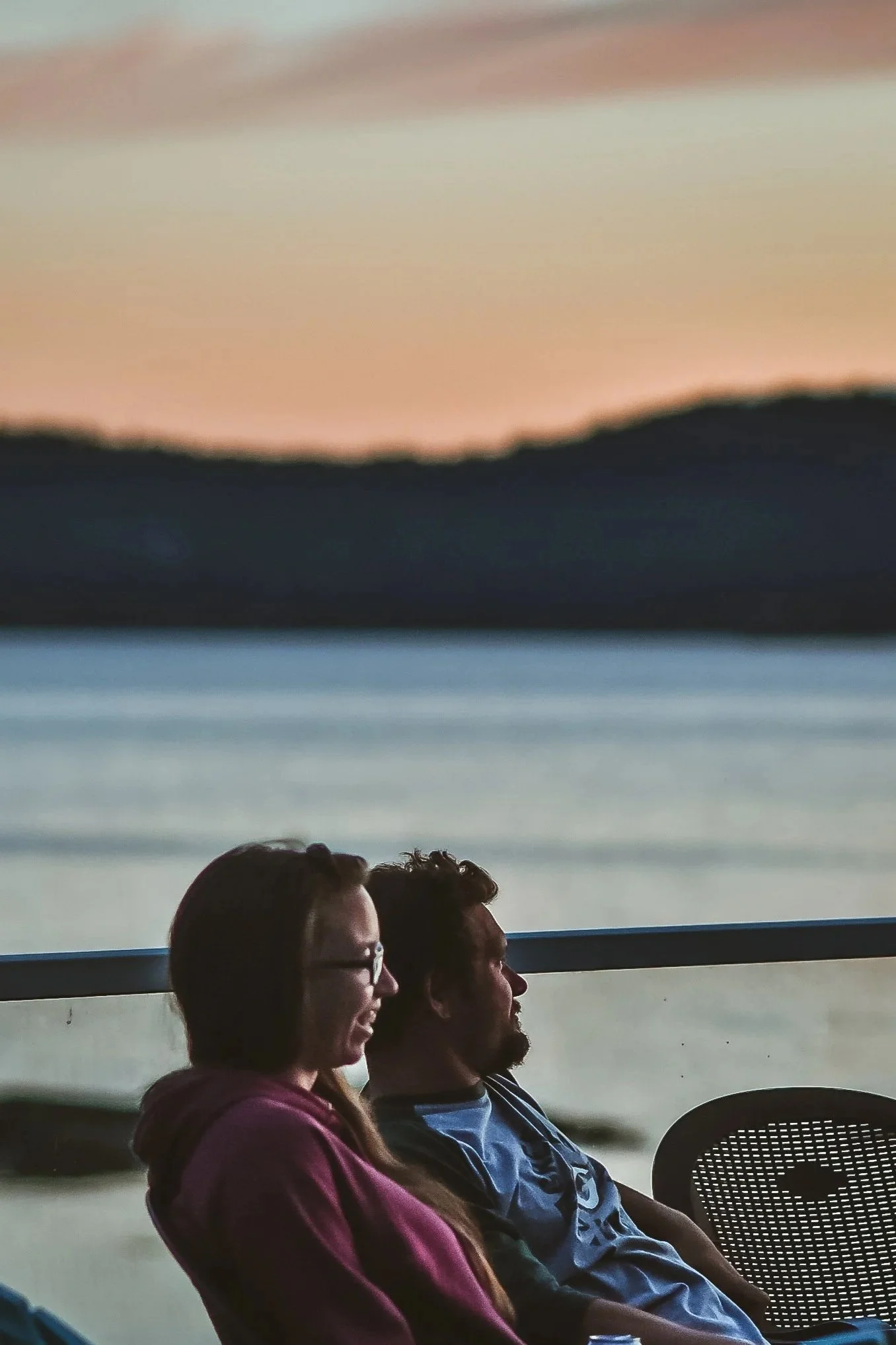 Two people sit outdoors near a body of water during sunset, facing to the right. The woman on the left is wearing glasses and a maroon hoodie, and the man on the right has a beard and is wearing a dark T-shirt. The background features a calm lake or river with dark hills and a colorful sky at dusk.