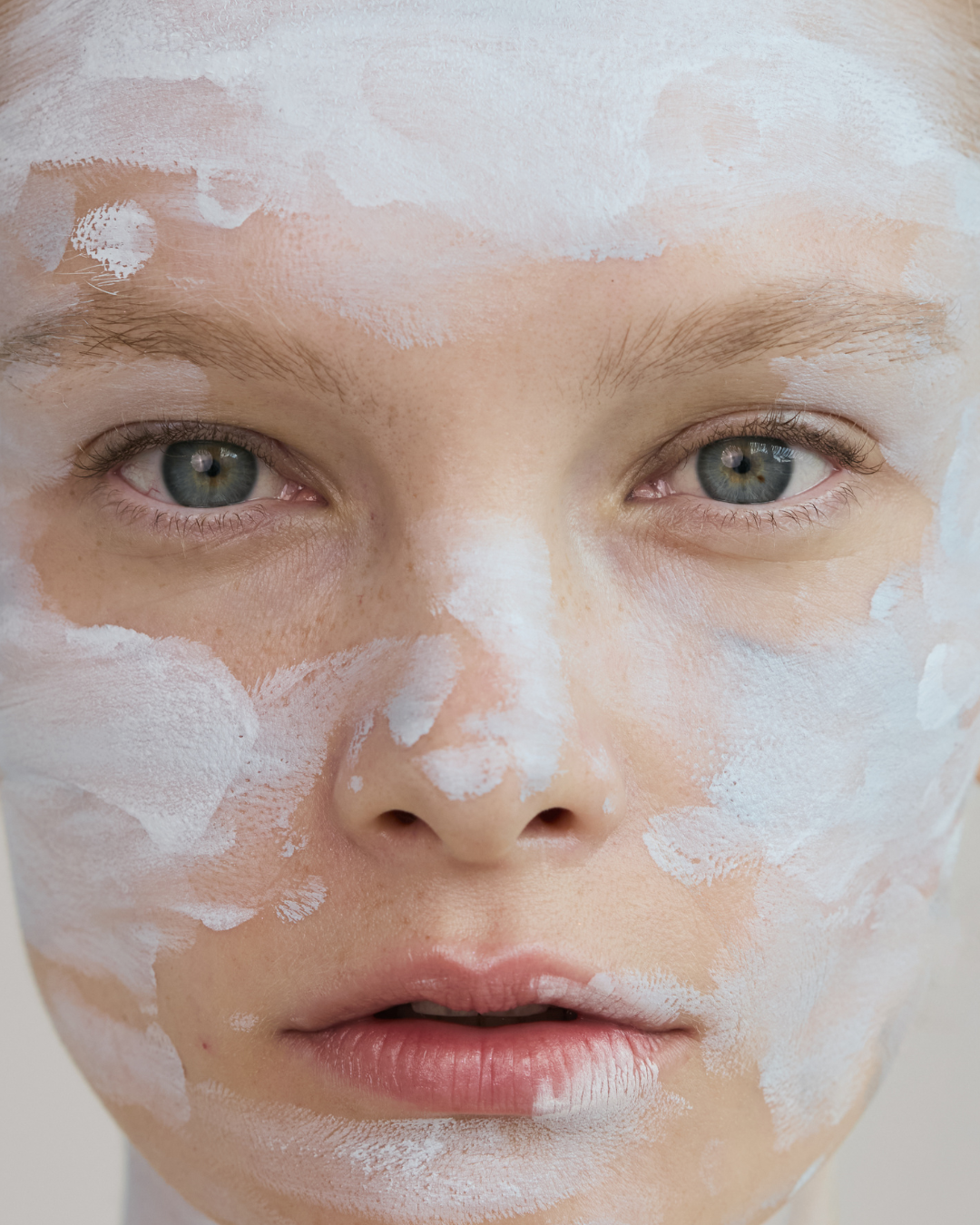 Close-up of a person's face with light skin and blue-gray eyes, covered in white facial mask or cream.