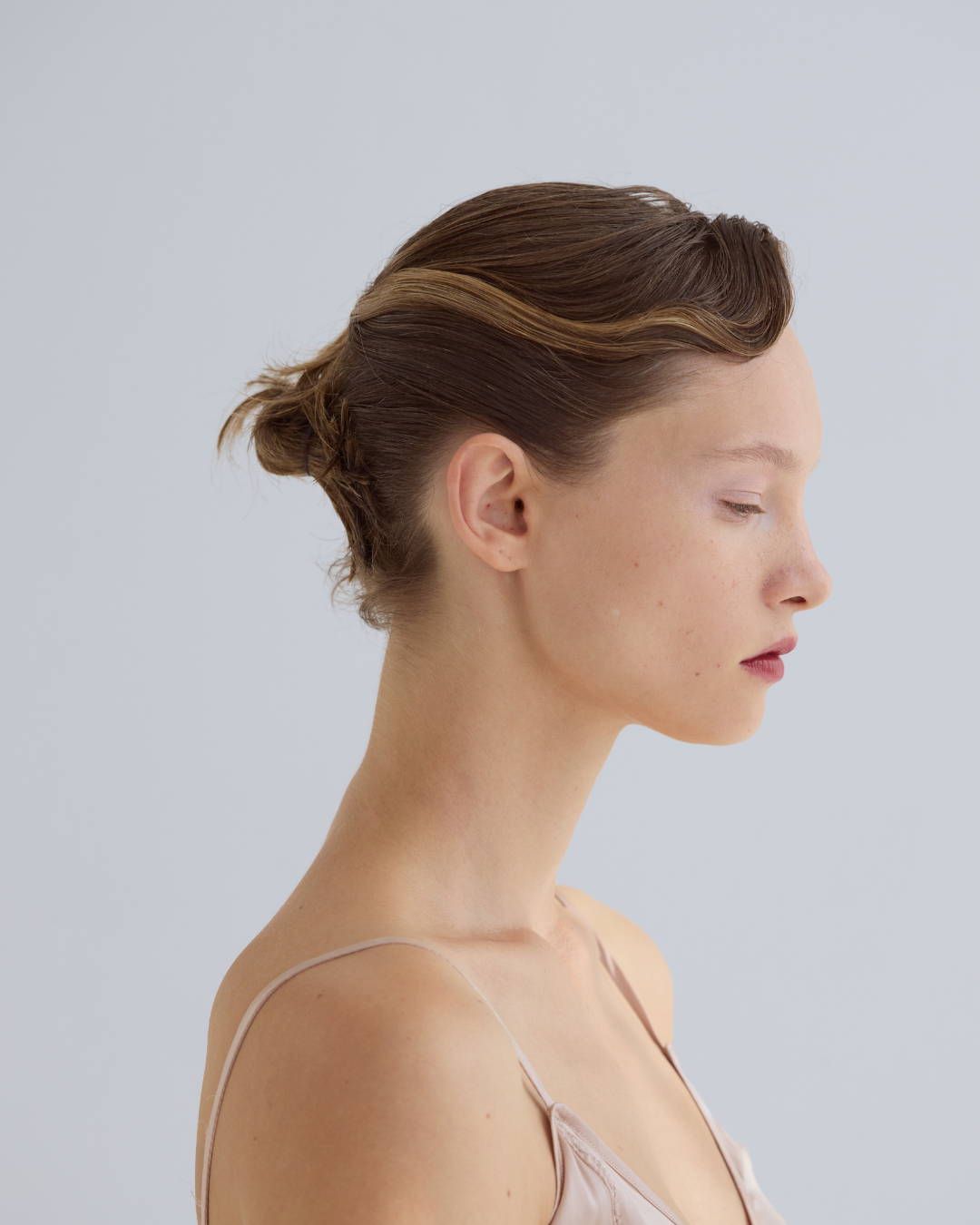 Profile of a woman with brown hair styled in a low bun, wearing a light-colored spaghetti strap top, against a plain light background.
