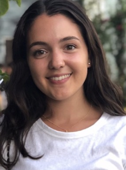 A young woman with long dark hair, smiling, wearing a white shirt and a delicate gold necklace, standing outdoors with greenery in the background.