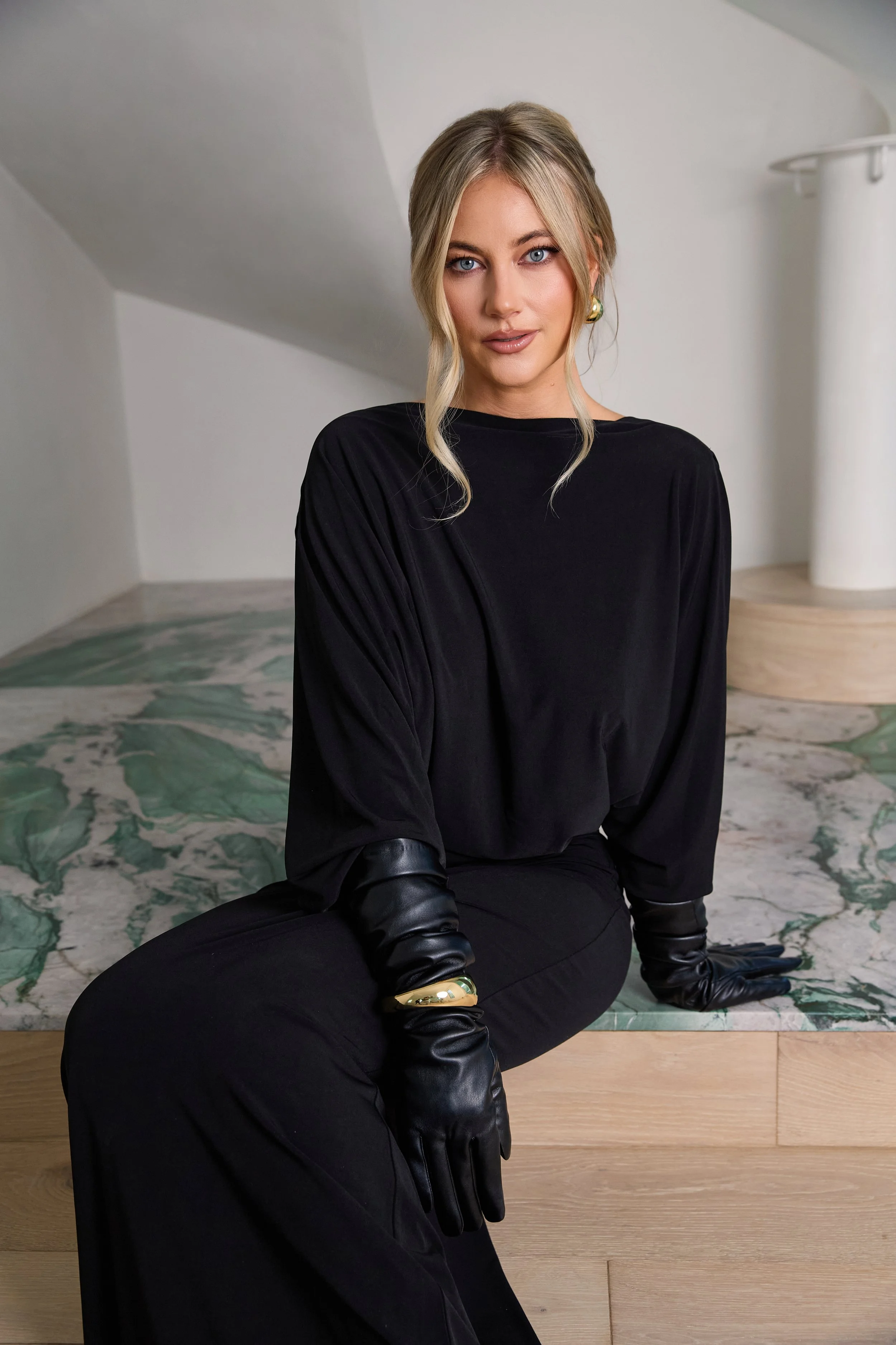 A blonde woman wearing a black long-sleeve top, black gloves, and matching black pants, sitting on a marble surface with a wooden accent, looking directly at the camera.