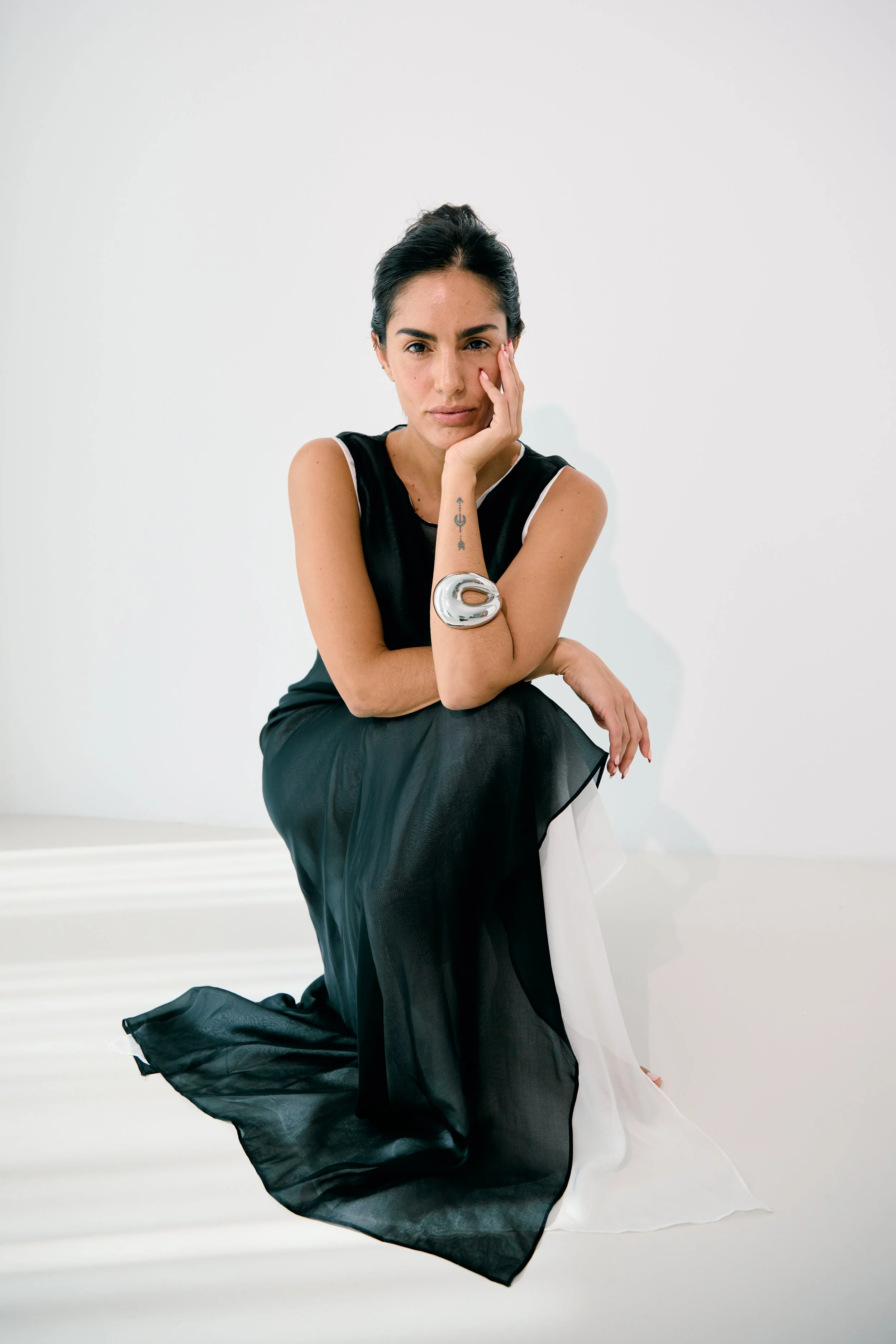 A woman with dark hair, wearing a black sleeveless dress with white accents, is sitting on a white surface against a plain white background. She has a silver bracelet on her left arm and a small tattoo on her inner left forearm. She has one hand resting on her face and is looking directly at the camera.