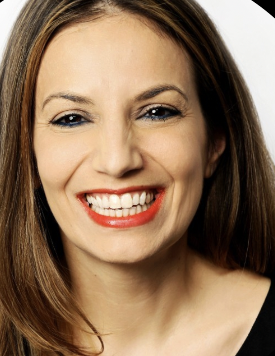 A woman smiling with red lipstick, showing her teeth, with long brown hair and a black top.