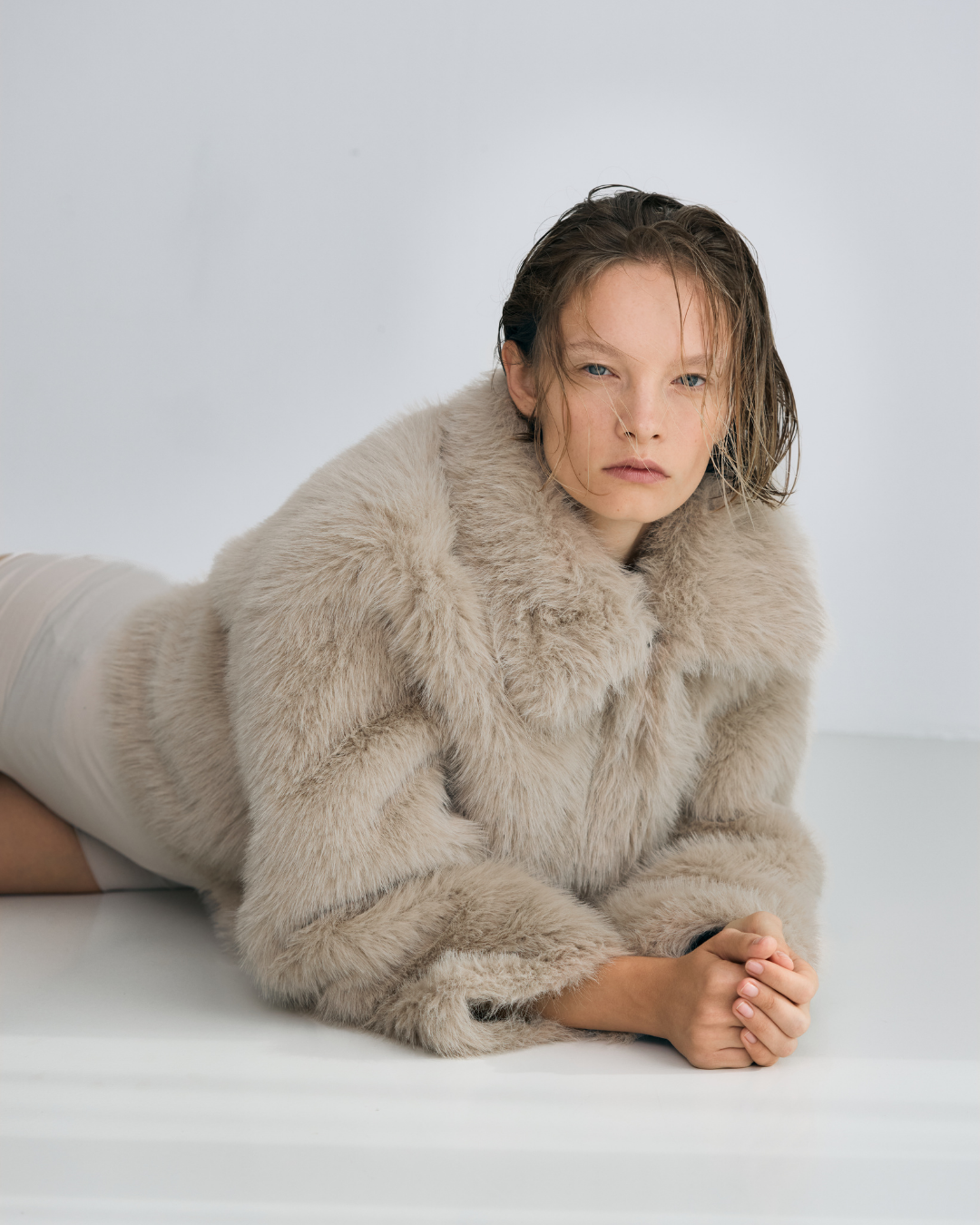 Woman lying on her stomach with hands clasped, wearing a beige faux fur coat, against a plain white background.