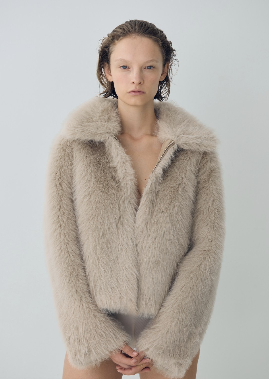 Young woman with short wavy hair wearing a beige fur coat, standing against a plain light background.