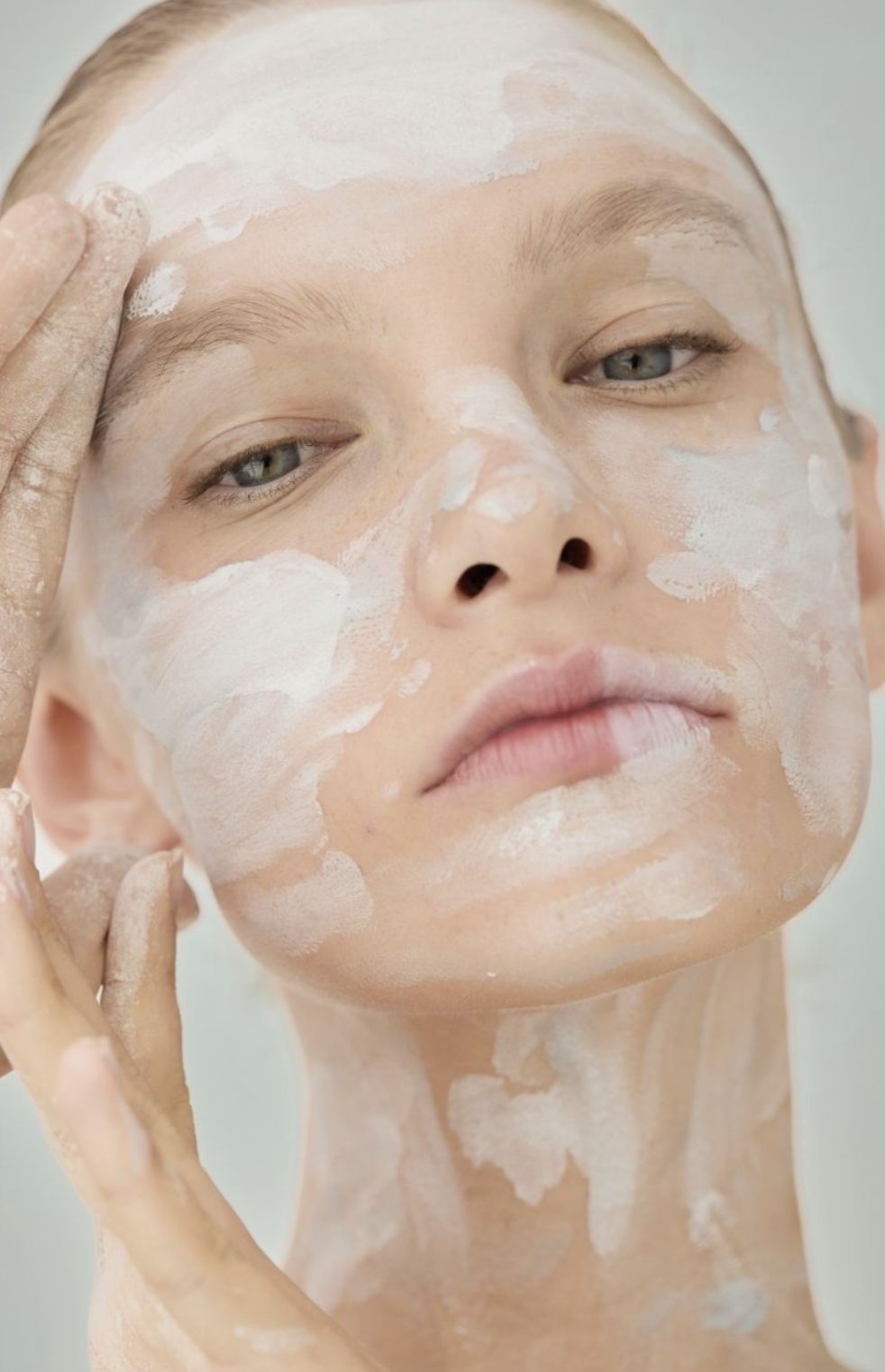A woman with light skin and blue eyes applying white face mask or skincare product to her face.
