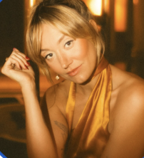 A young woman with short blonde hair and a yellow dress, looking at the camera with a slight smile in a dimly lit setting.