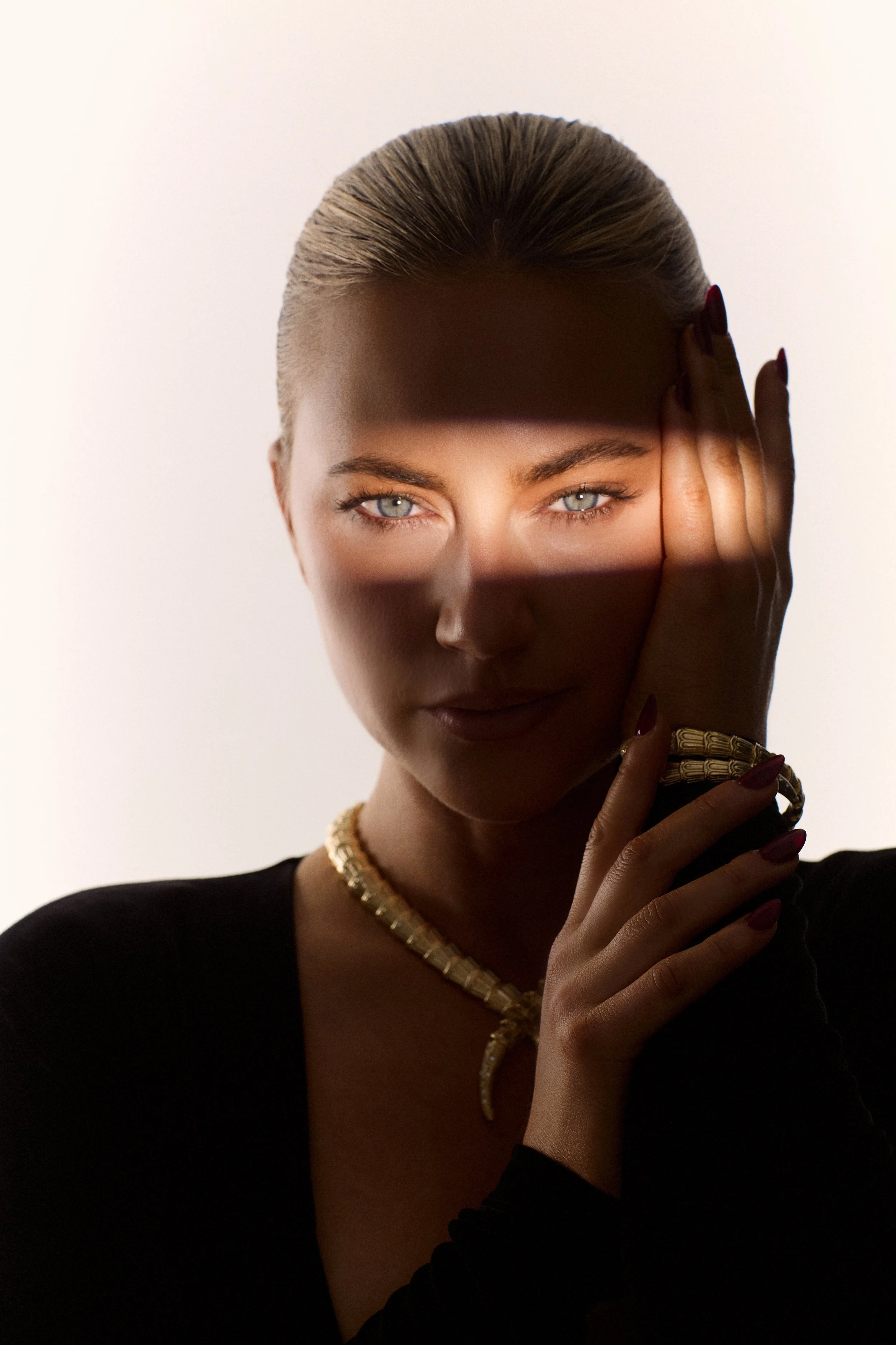 A woman with blonde hair and blue eyes wearing a black top, gold necklace, and bracelet. She is posing with her right hand on the side of her face, partially covering her face with her hand, with a shadow crossing her face from a horizontal object.