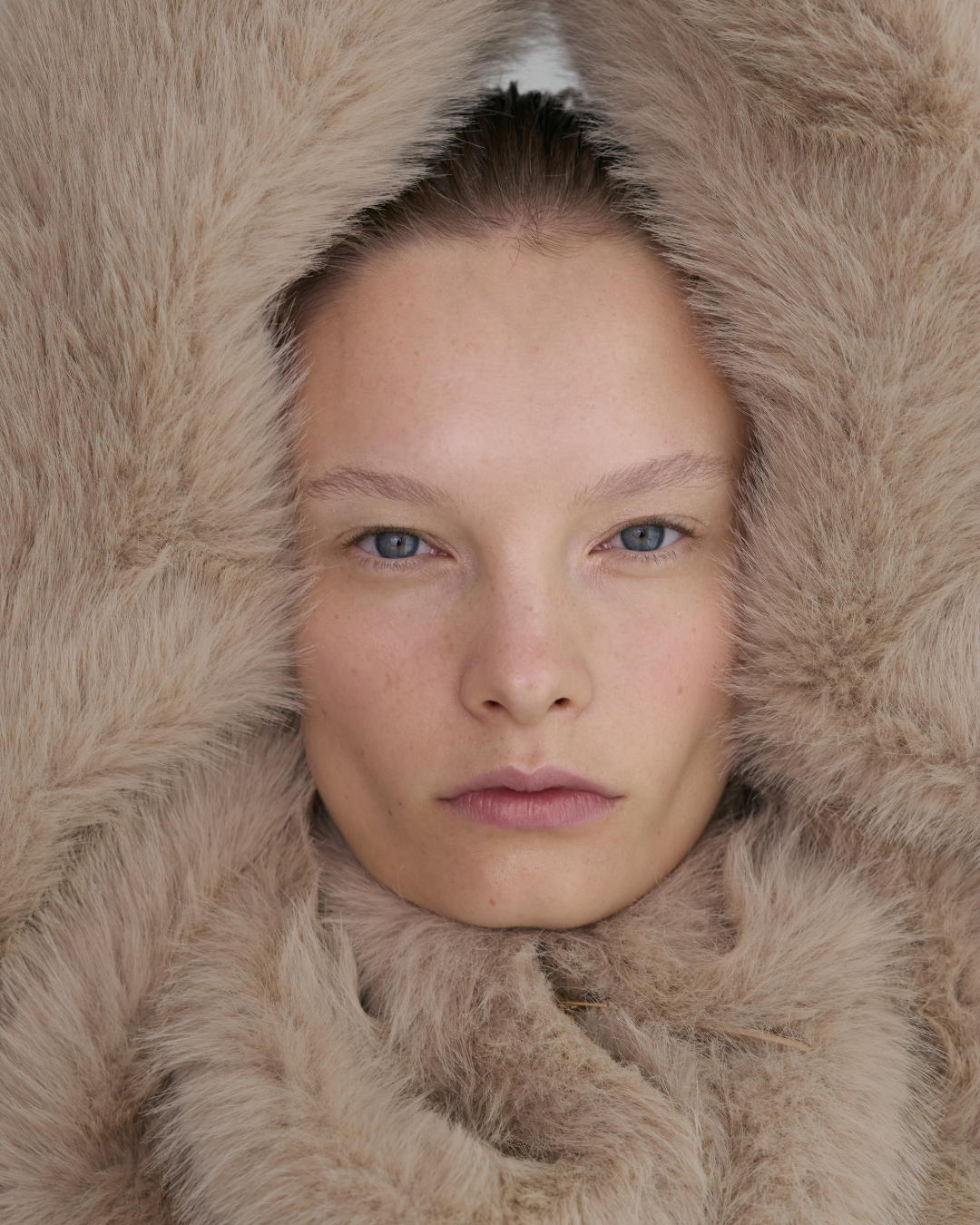 Close-up of a young woman with blue eyes and freckles, surrounded by a large, fluffy, beige fur.