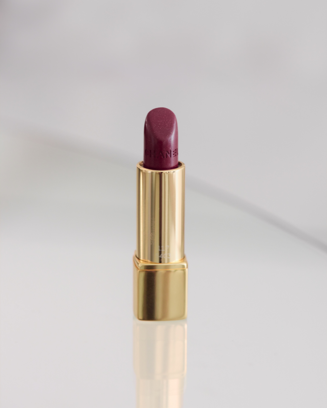 A tube of burgundy lipstick with a gold case, standing upright on a reflective surface against a plain light background.
