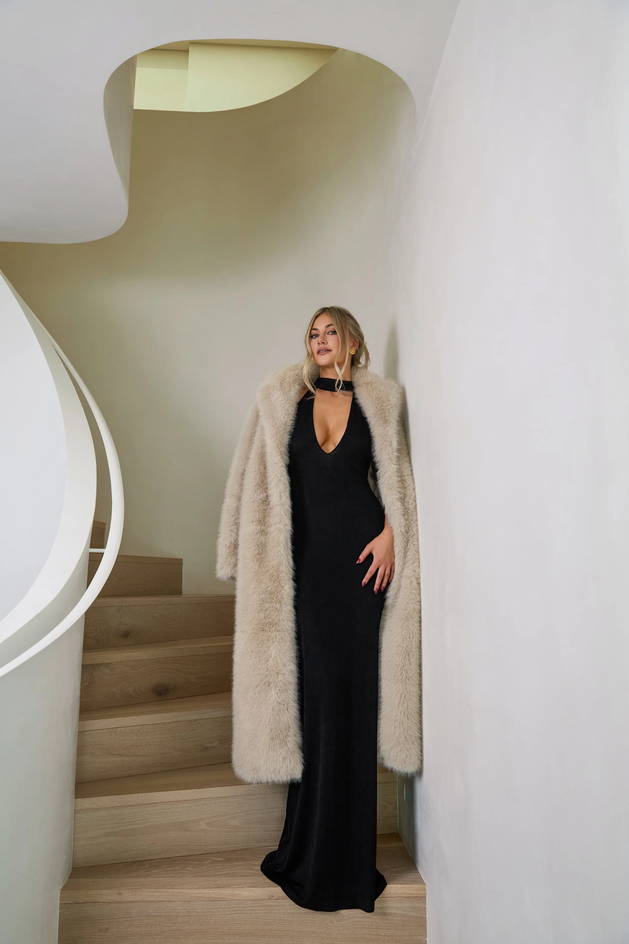 A woman standing on a wooden staircase in a modern interior, wearing a black dress and a long beige fur coat.