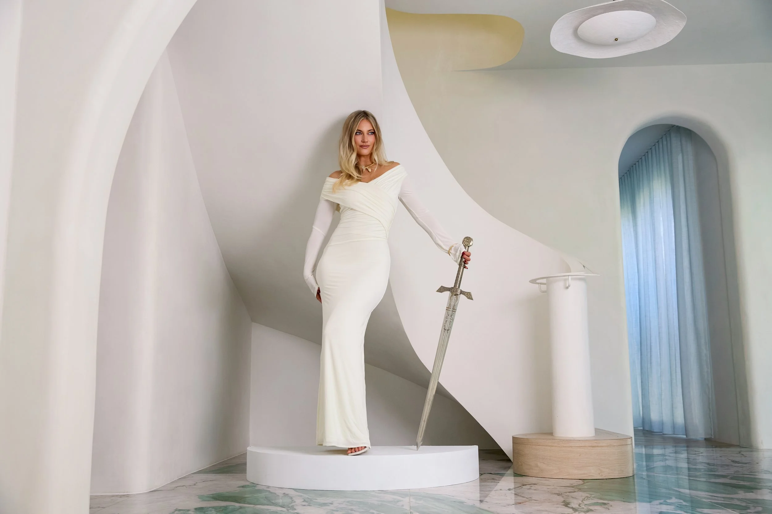A woman with long blonde hair wearing an elegant white gown and long white gloves stands on a round white pedestal. She holds a large sword with an ornate hilt in her right hand. The background features modern white architectural elements with curved walls and a column, along with a window with sheer curtains.