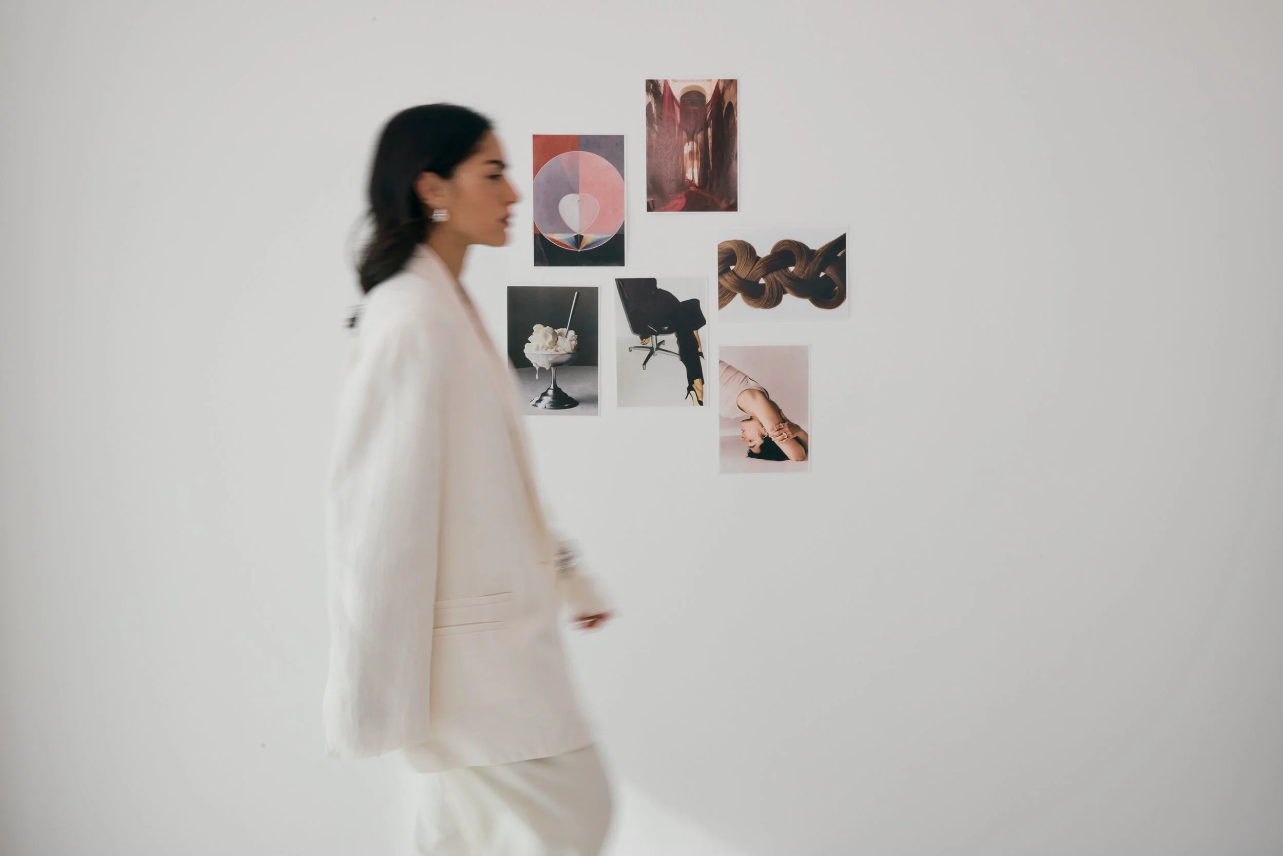 A woman in a white suit walking past a gallery wall with colored abstract and artistic photographs and prints.