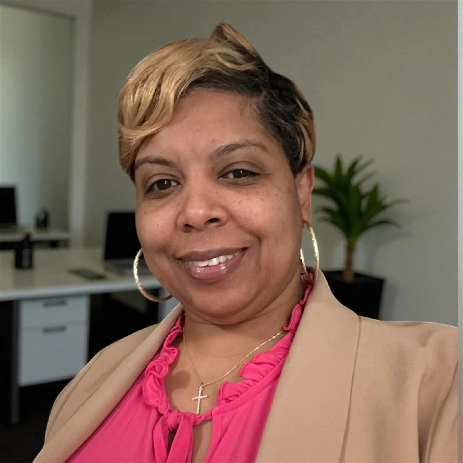 A woman with short, styled hair smiling at the camera, wearing large hoop earrings, a pink ruffled top, a beige blazer, and a cross necklace inside an office with computers and a potted plant in the background.