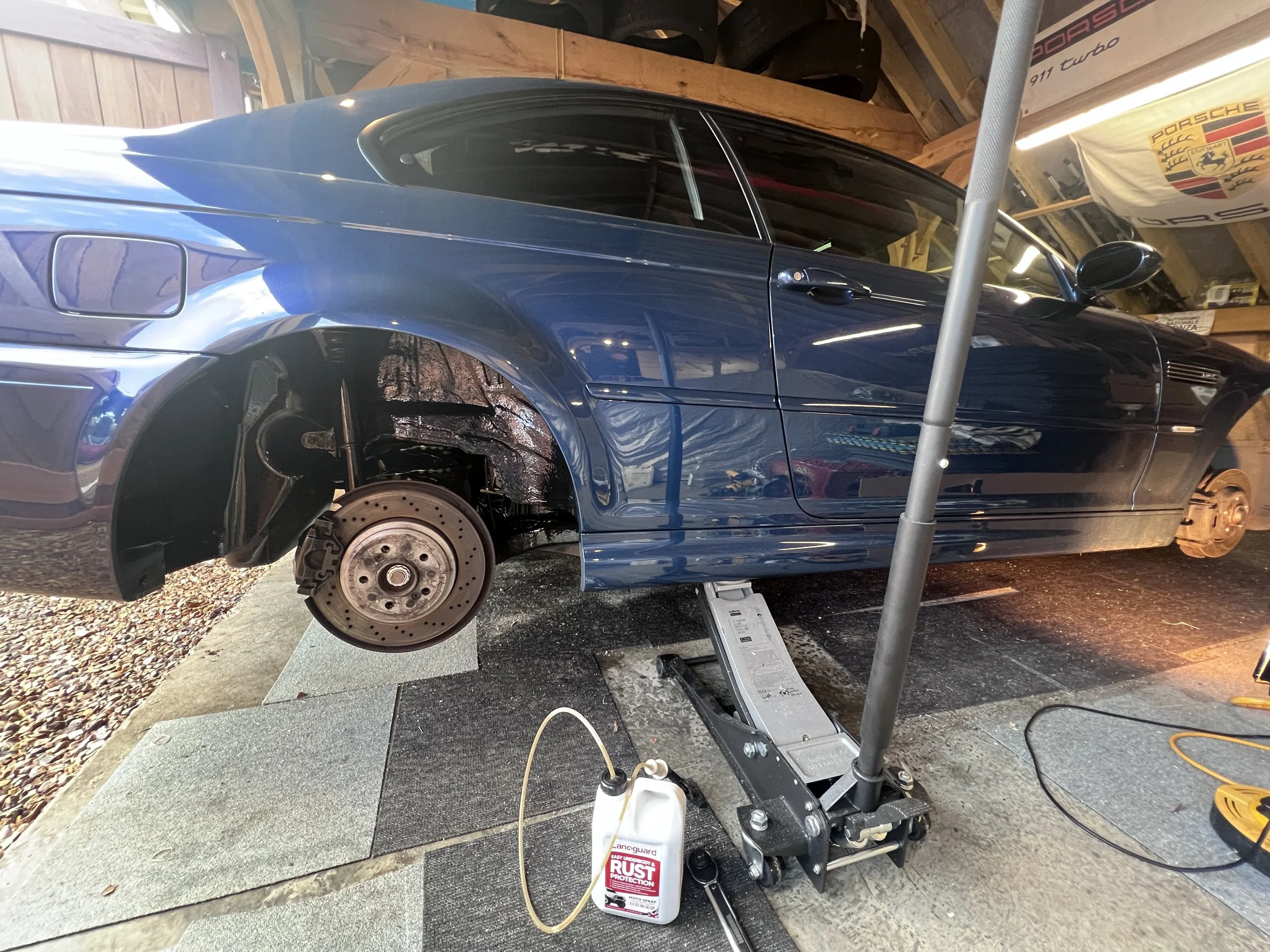 A blue sports car with the front wheel removed, lifted on a floor jack in a garage with Porsche banners hanging on the wall.