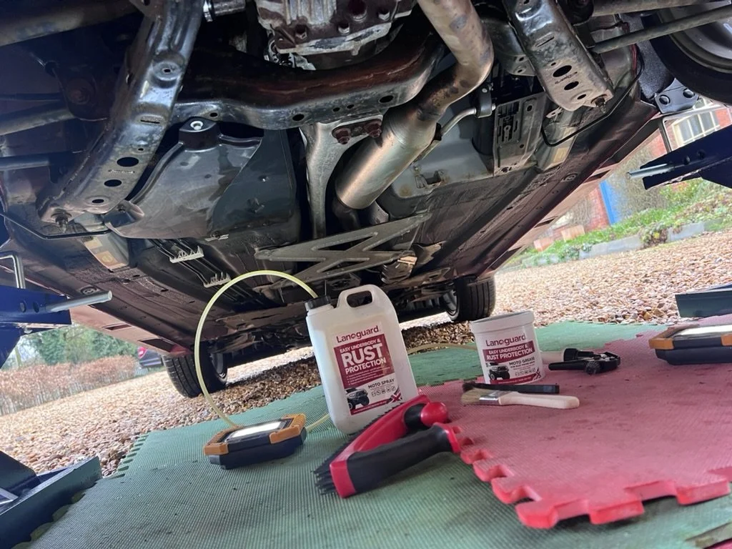 Underneath view of a car in a garage with tools and rust protection products on the floor.