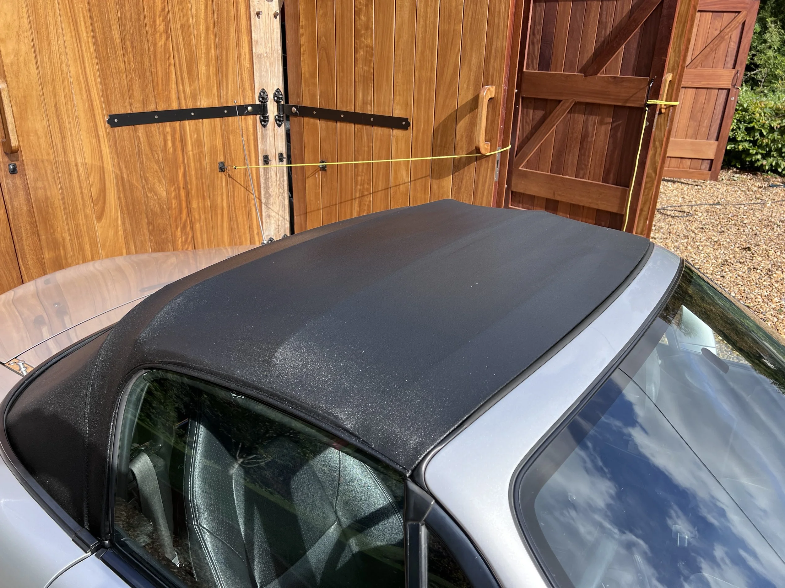 The image shows a silver convertible car with a black soft top parked in front of a wooden fence and gate. The gate is partially open and secured with yellow strings tied to nearby hooks.