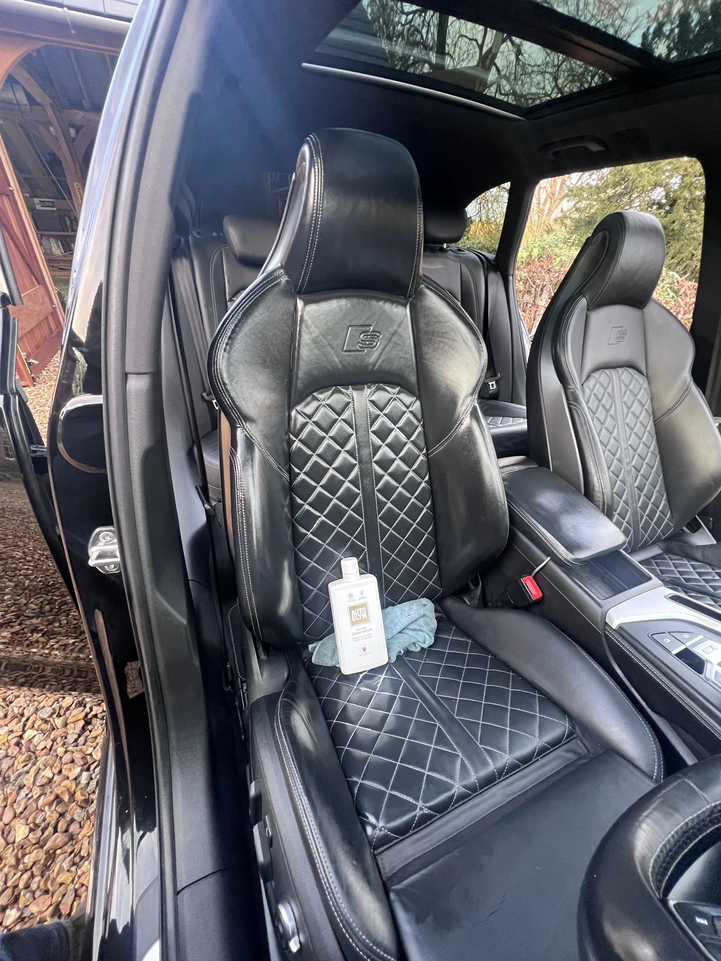 The image shows the front seats of a black car, with the driver's seat in the foreground. The seats are black leather with diamond-pattern stitching. There is a bottle of hand sanitizer, a cleaning cloth, and a small container on the driver's seat.