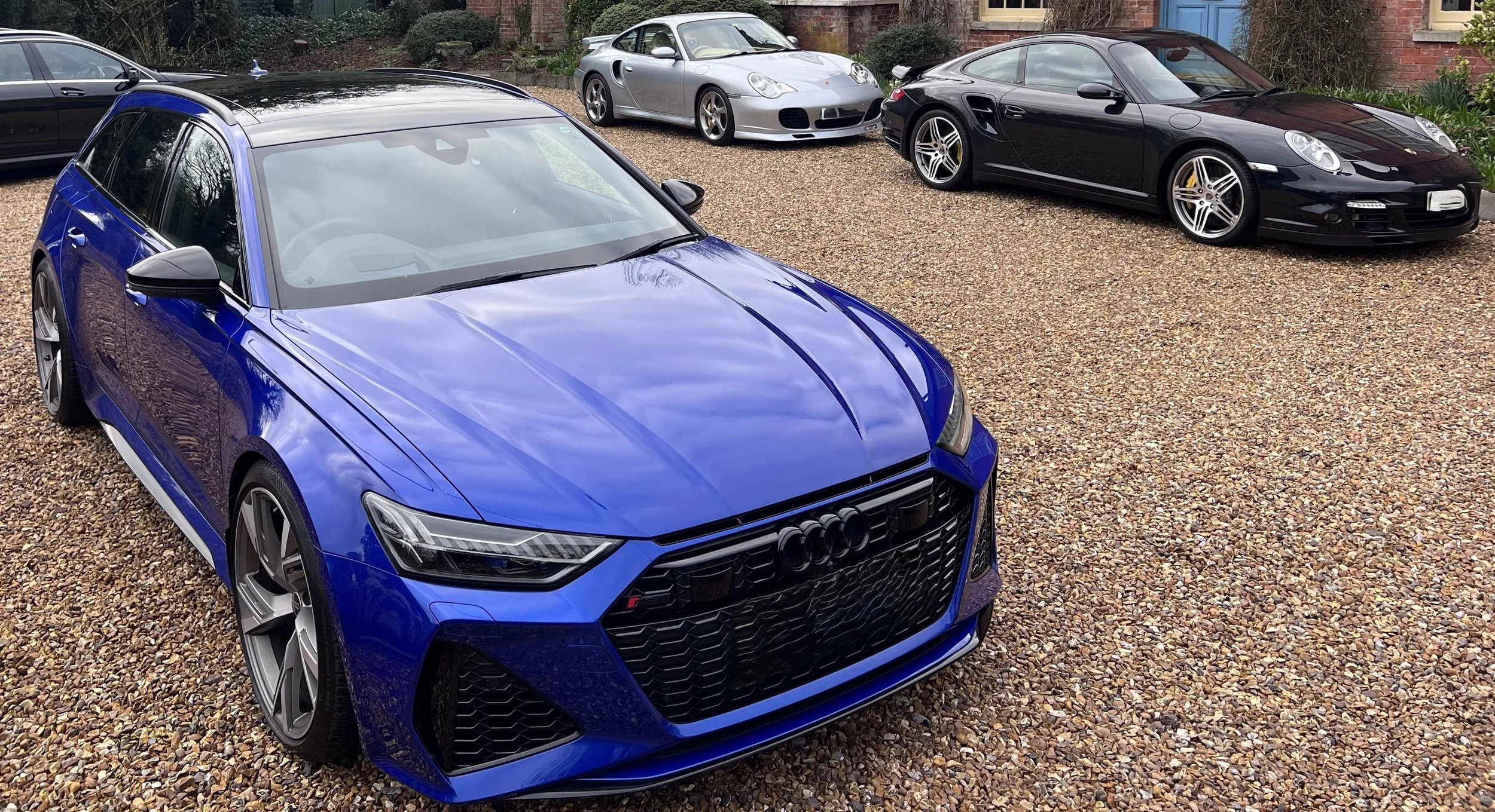 A blue Audi car in the foreground with a black car and three Porsche cars parked behind on a gravel driveway outside a house.