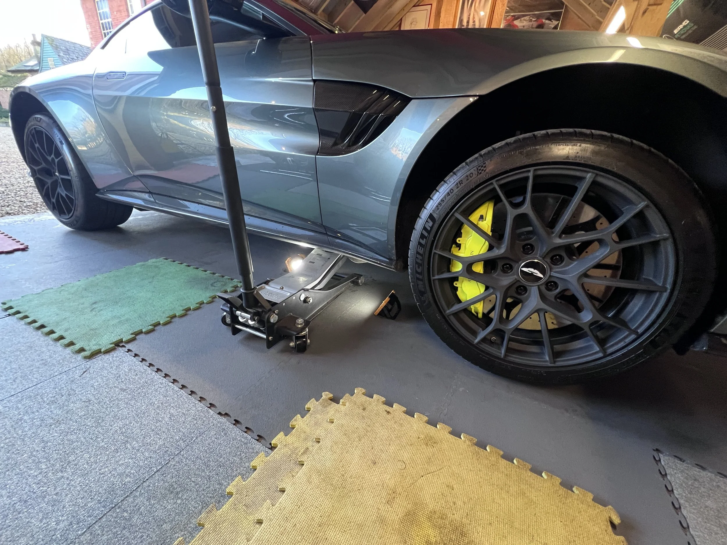 A blue sports car with yellow brake calipers on a floor with colored puzzle mats, raised on a hydraulic lift inside a garage.