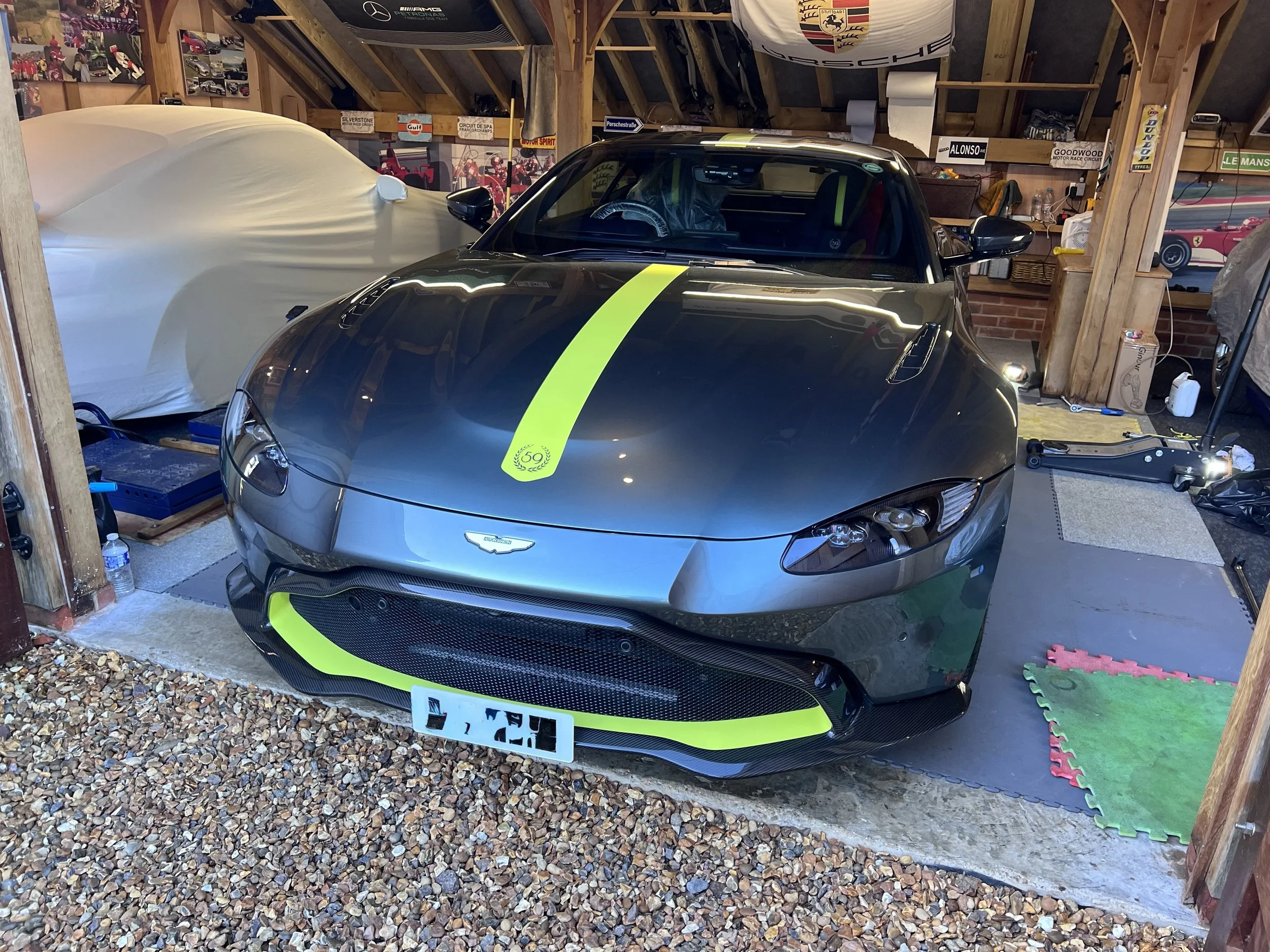 A grey sports car with yellow racing stripes parked inside a garage. The garage has various items including a covered object on the left, car posters on the walls, and a small scooter on the right. The car has a black grille with green accents.