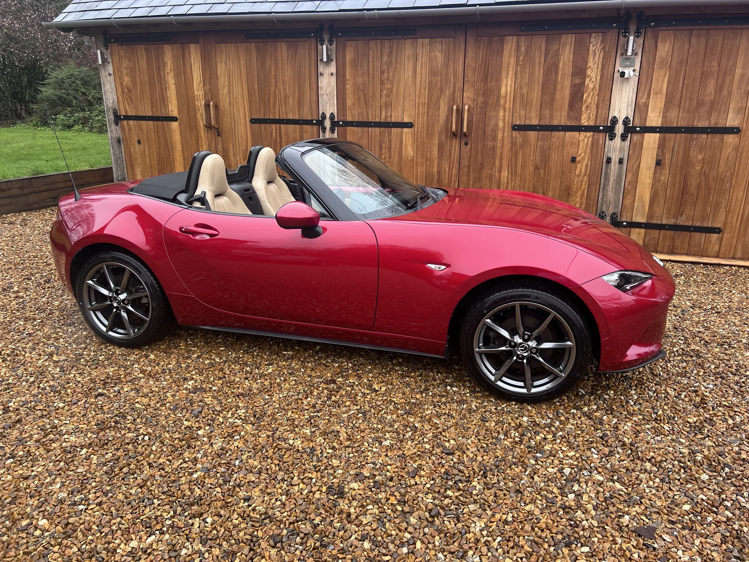 Red convertible sports car parked on gravel driveway with a wooden garage door in the background.