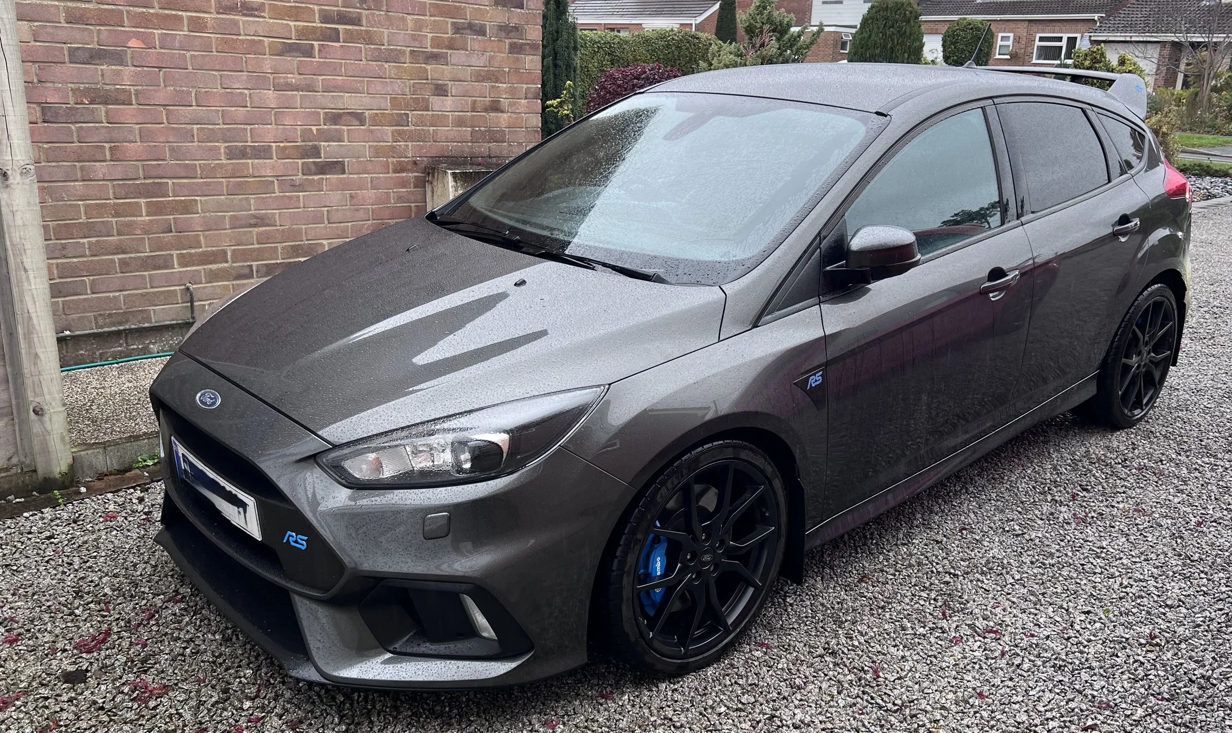 Gray Ford Focus RS parked on gravel driveway next to a brick wall with houses in the background, showing raindrops on the car's surface.