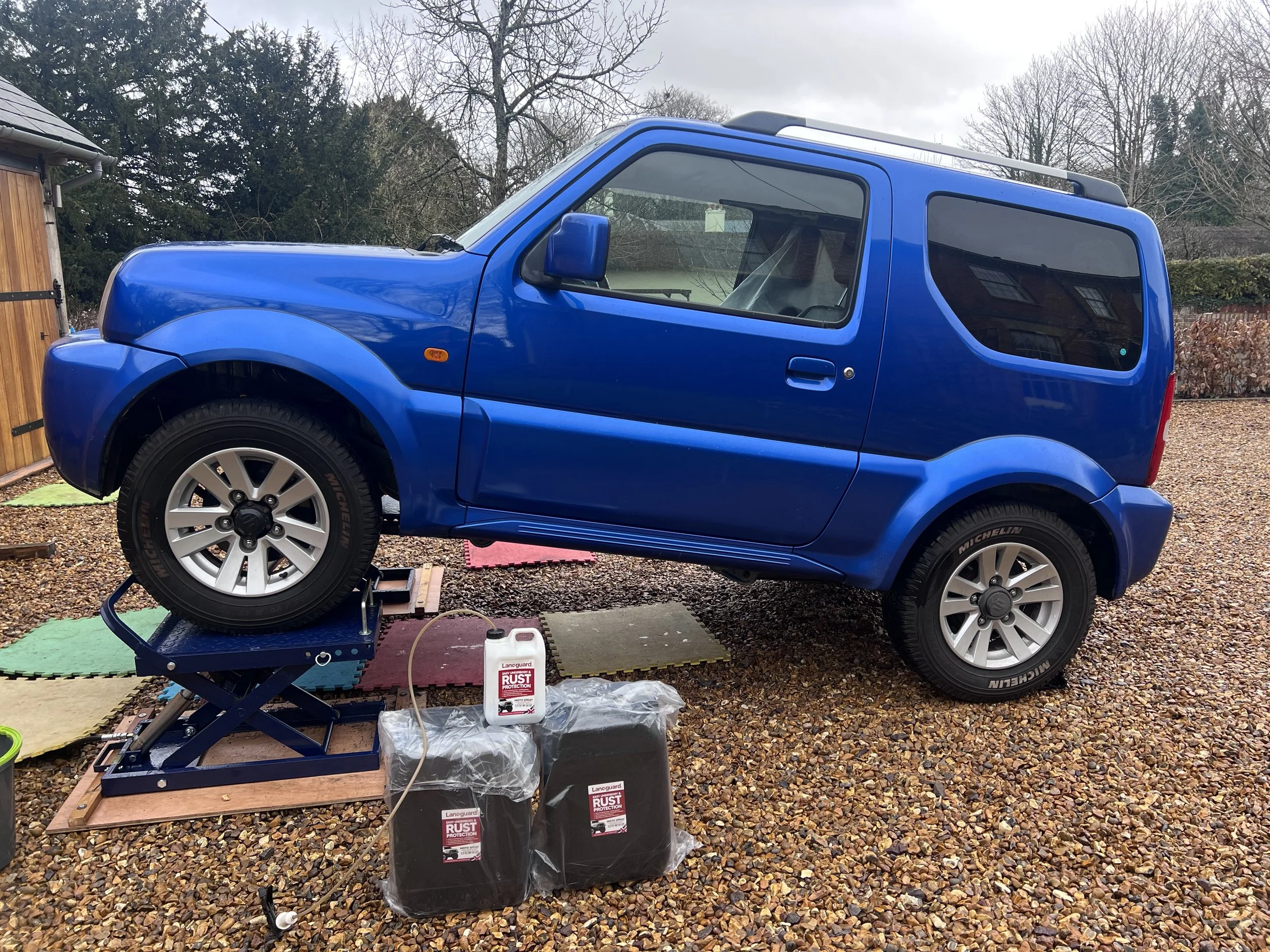 A blue car is lifted on a hydraulic platform outside in a gravel area, with car maintenance supplies around it, including containers of rust protection liquid and foam mats on the ground.