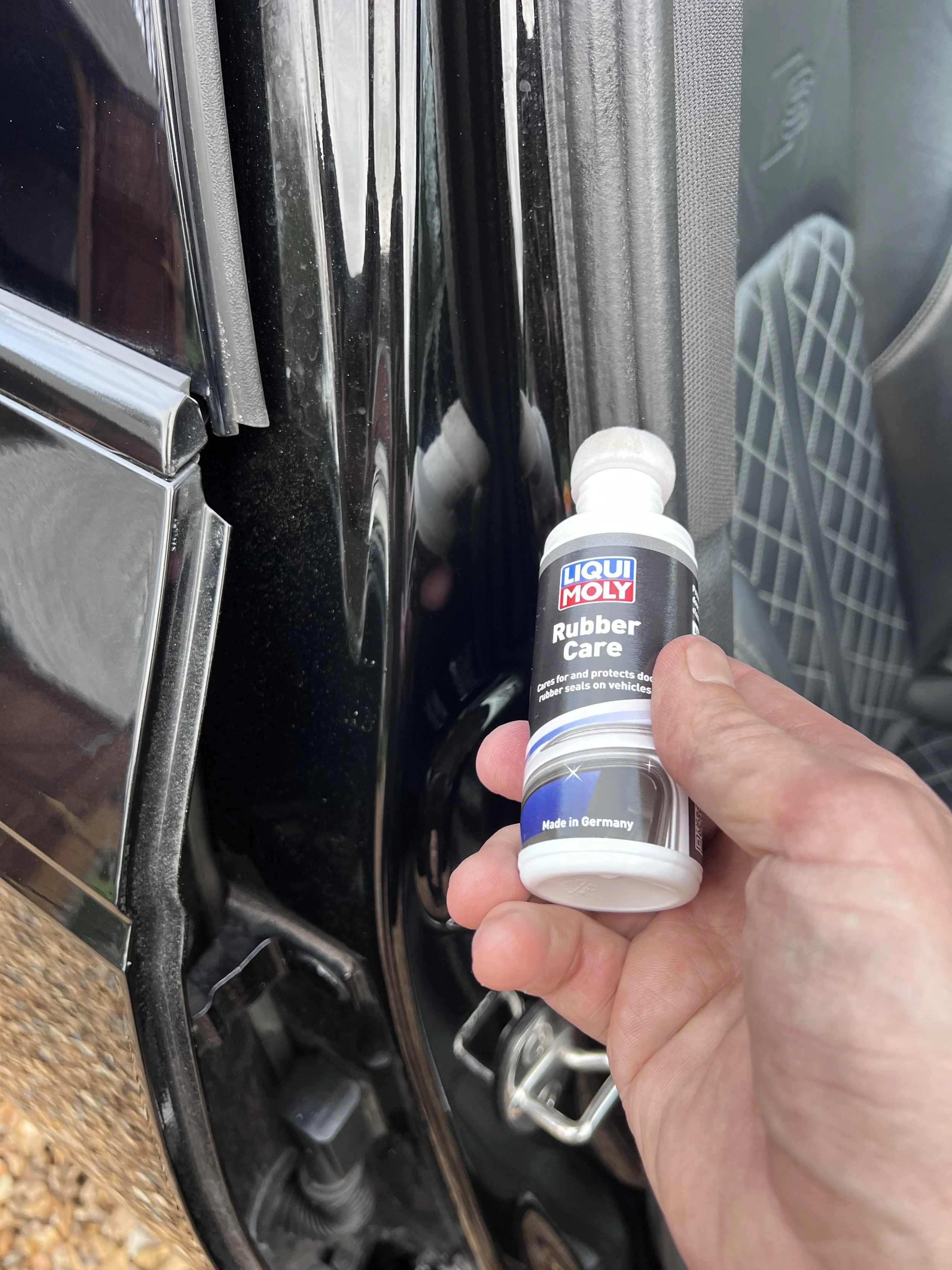 Person holding a small bottle of Liqui Moly rubber care in front of a black car door to apply protection and sealing for door rubber seals.