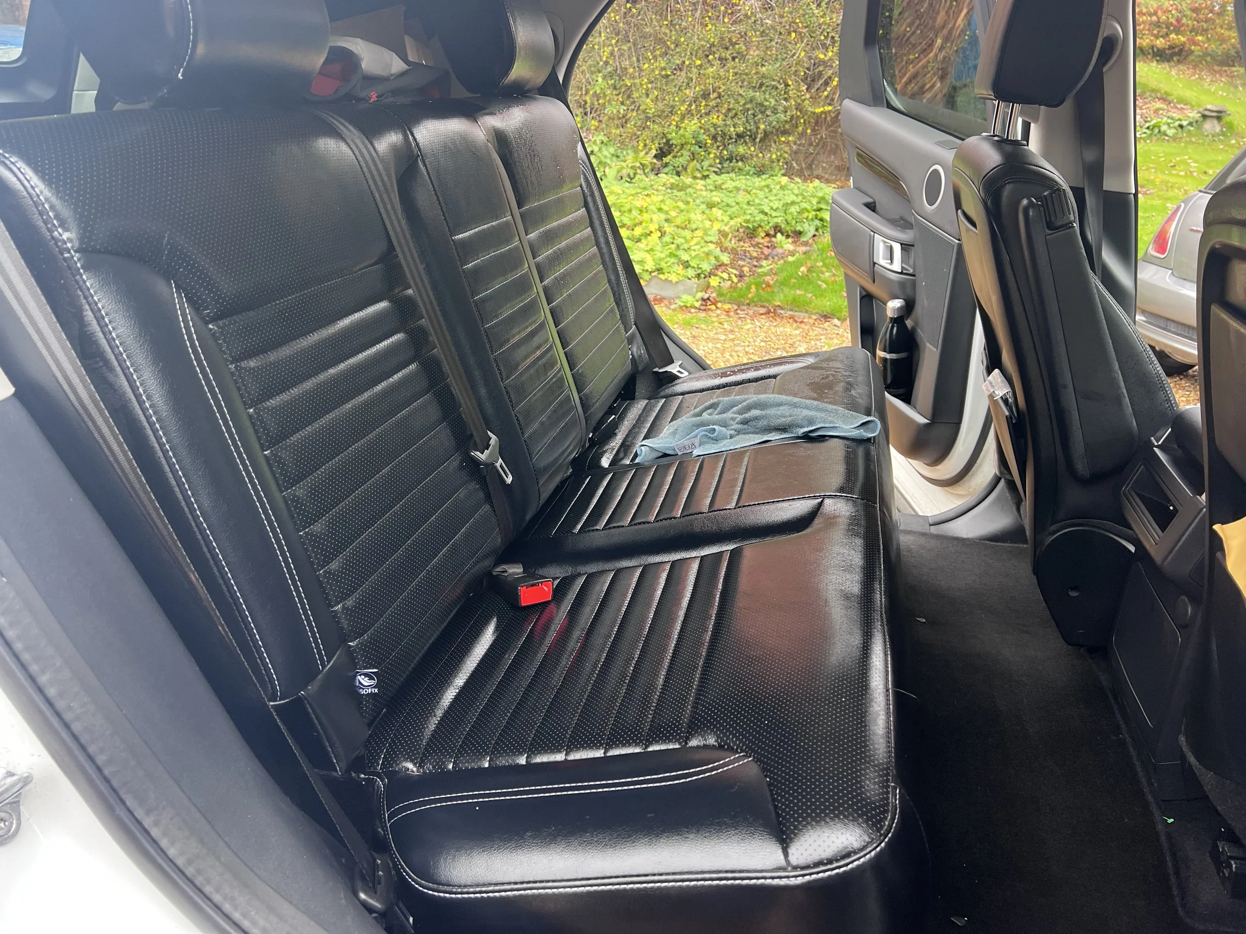The interior of a vehicle showing a back seat with black leather seats, a cloth on the seat, and outside greenery visible through the windows.
