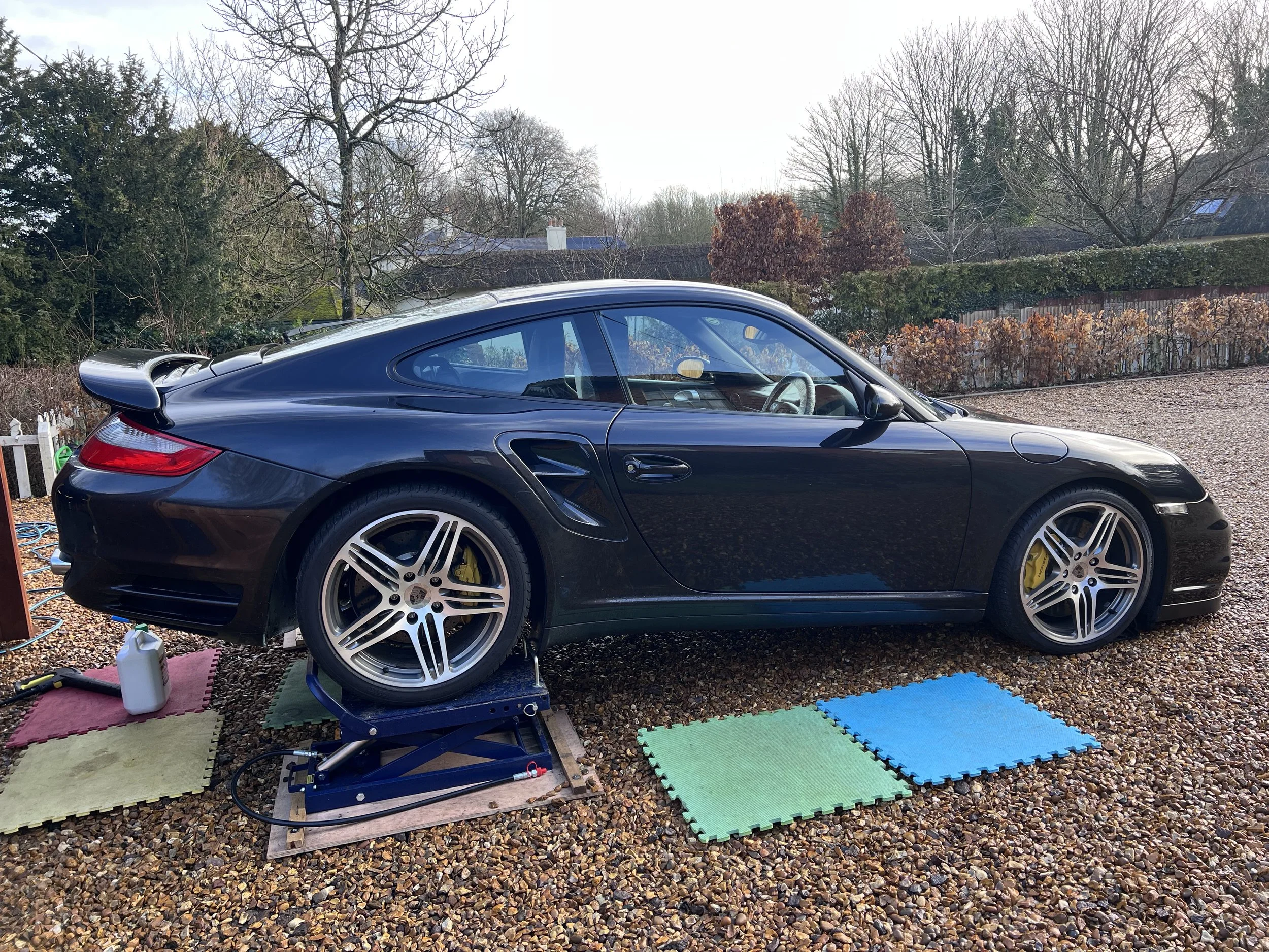 Black Porsche sports car parked outdoors on a gravel driveway, with repair tools, mats, and a hydraulic lift nearby.