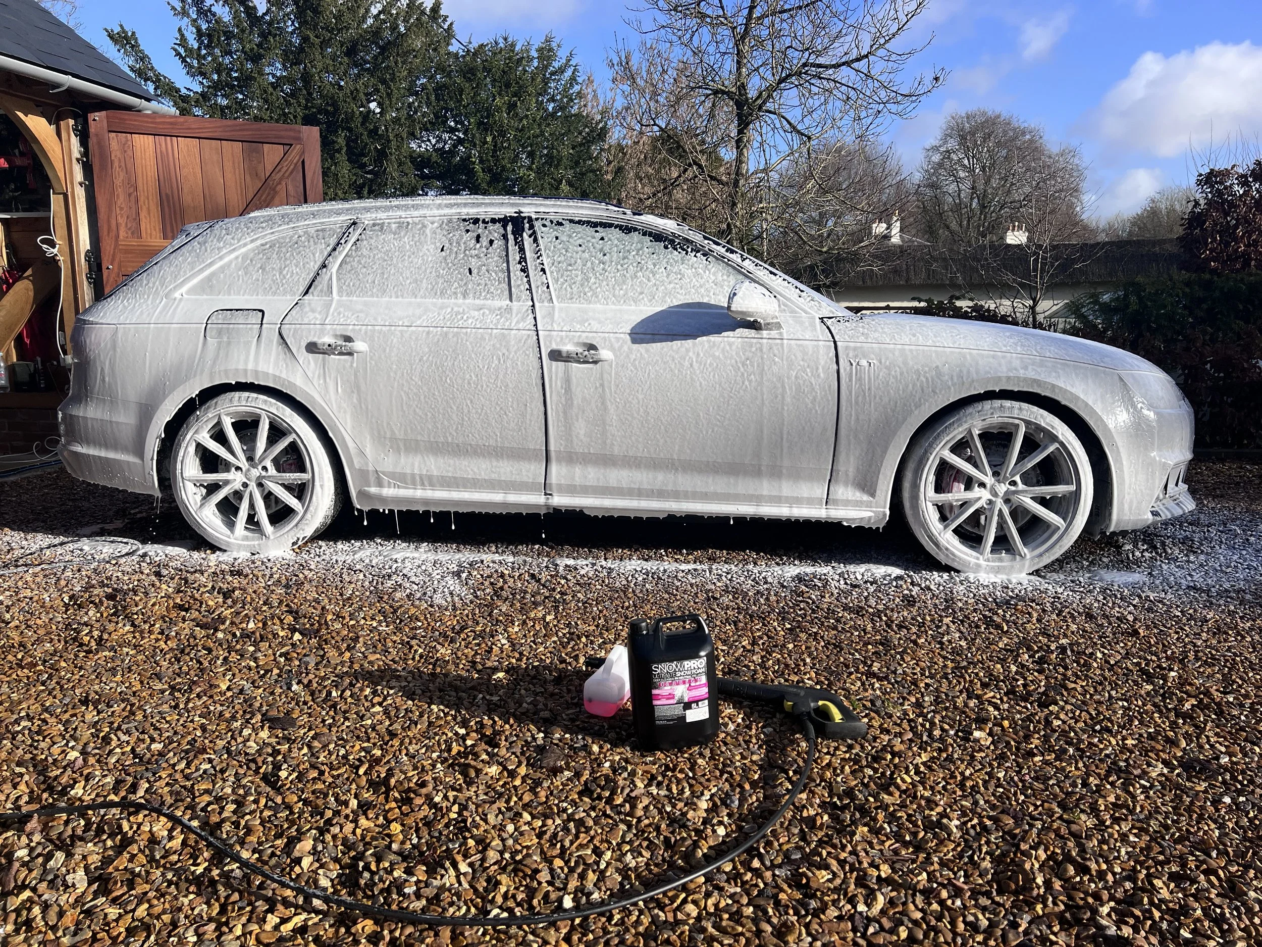 A silver car being washed with foam and soap, parked on a gravel driveway, with a garden shed, trees, and a cloudy sky in the background.