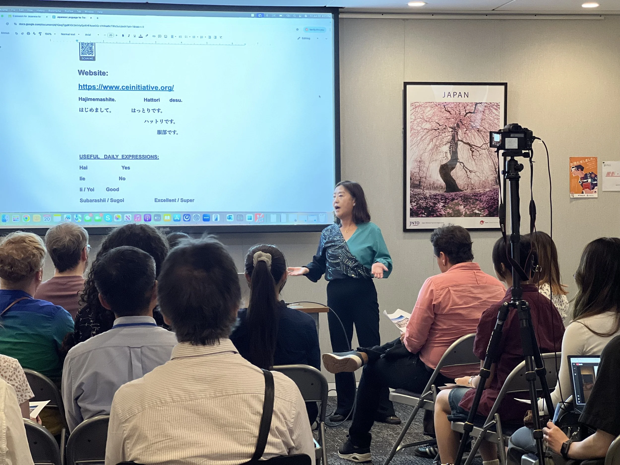 A woman is speaking to an audience in a conference room. There is a large screen behind her displaying a webpage with some Japanese text and a QR code. An audience is seated and listening, and there is a camera set up in front of the woman. There is a poster on the wall with a cherry blossom tree and the text 'JAPAN'.