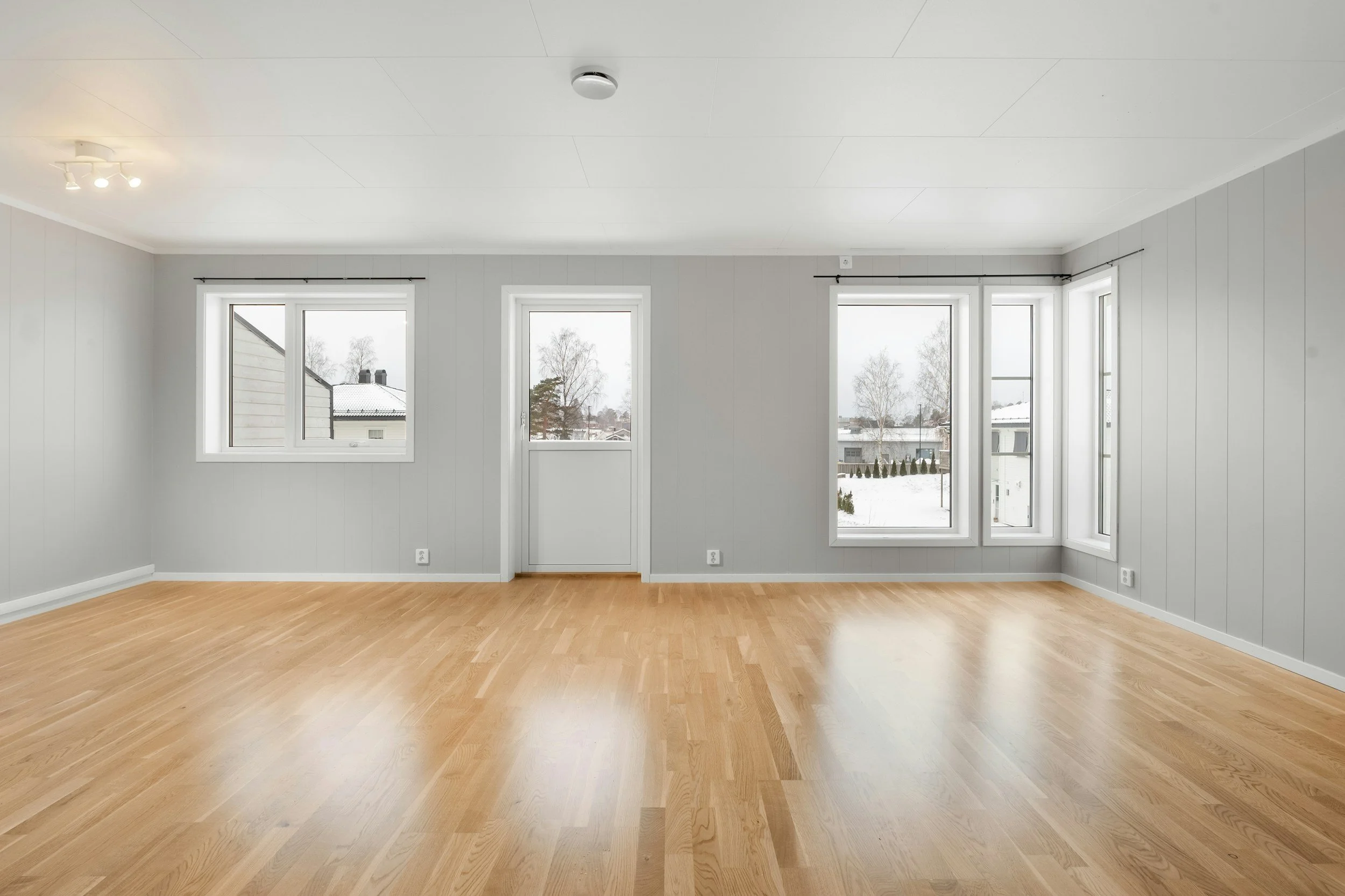 Empty room with white walls, large windows, wooden flooring, and ceiling lights showcasing a winter landscape outside.