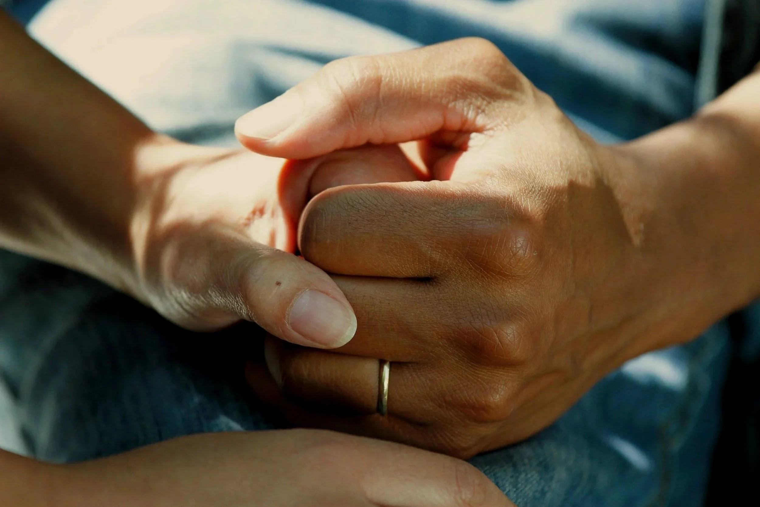 Two people holding hands, one with a light skin tone and the other with a darker skin tone, wearing a wedding band.