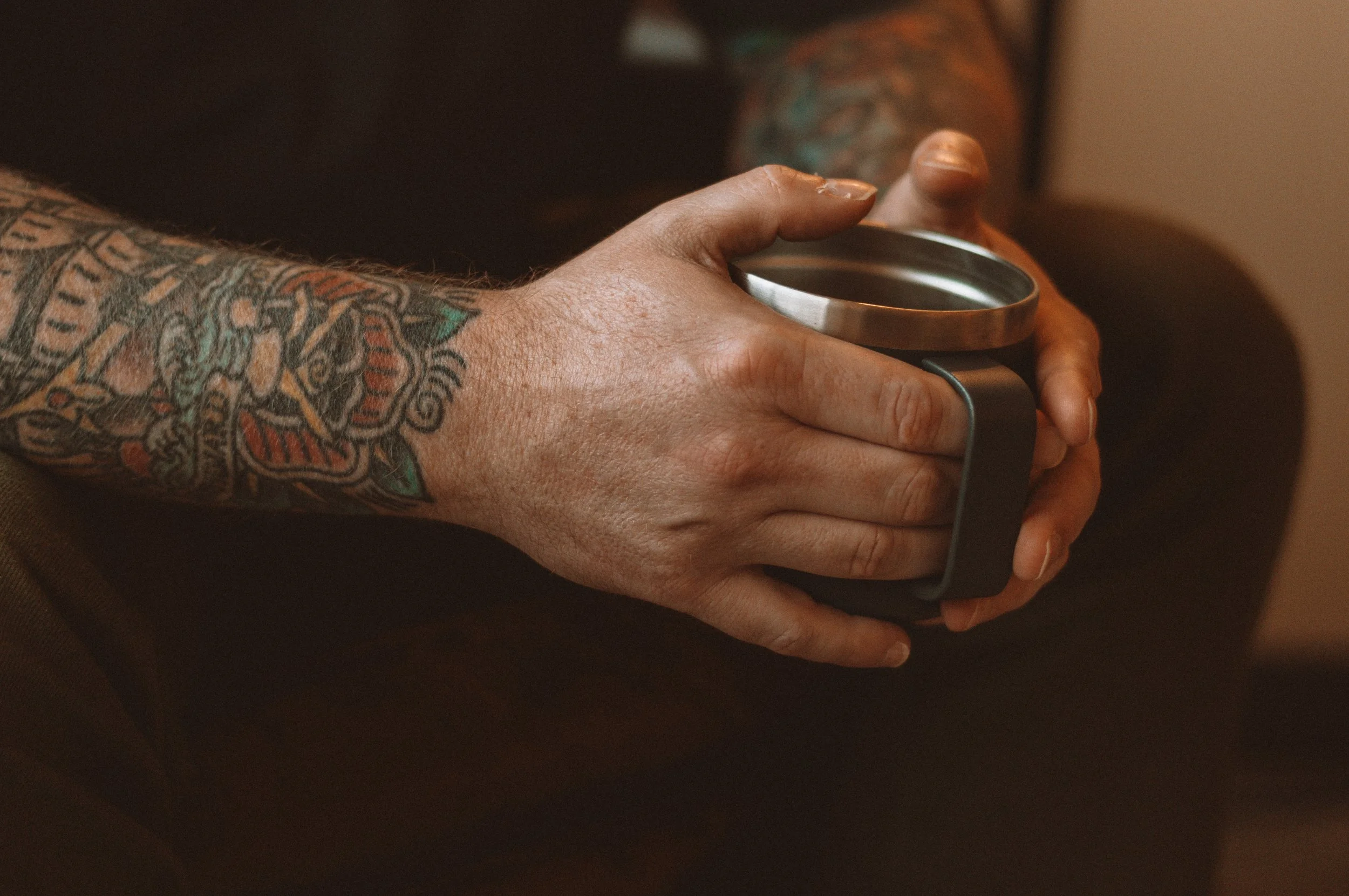 A tattooed man holding a stainless steel mug with both hands, sitting on a couch.