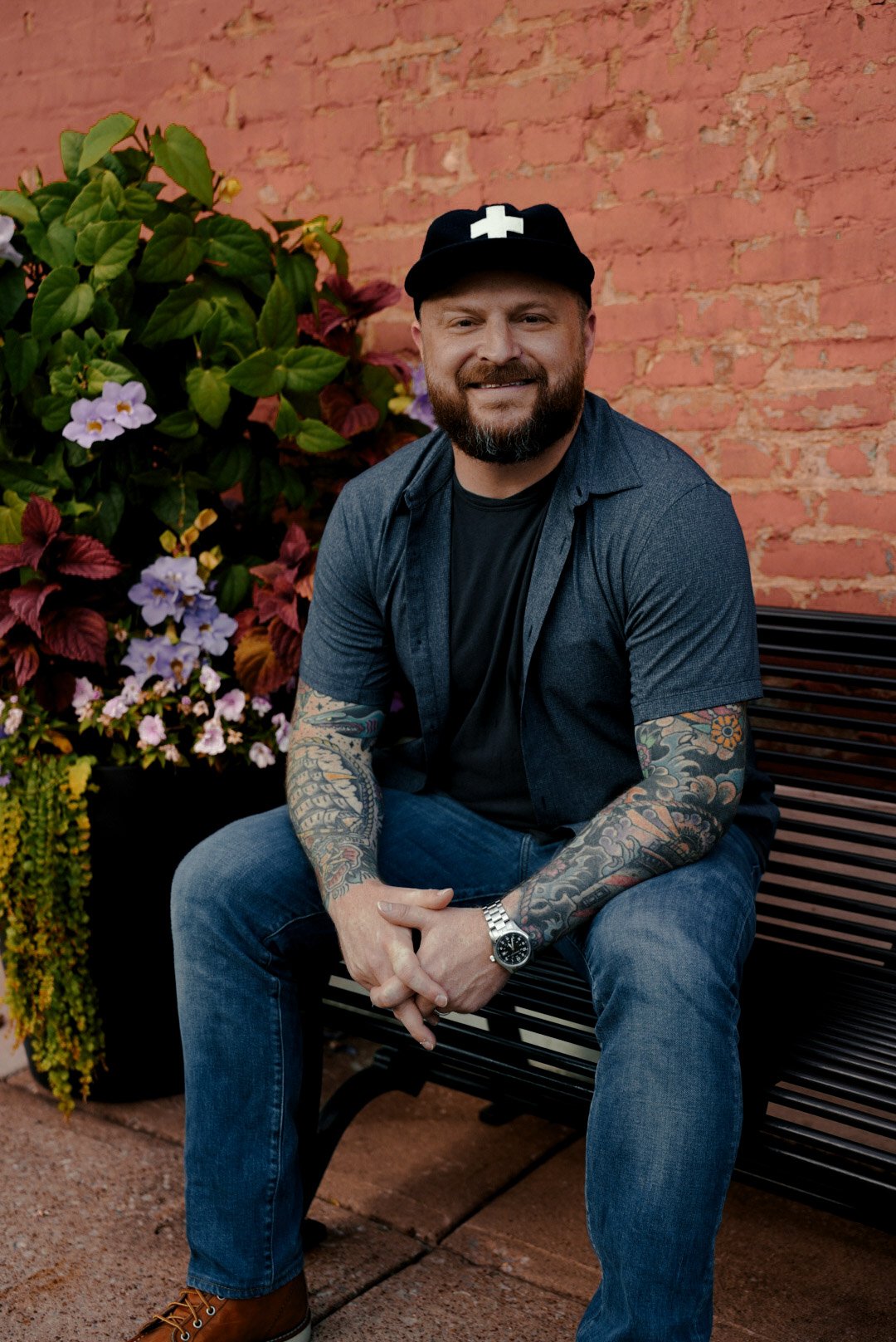A man with a beard and tattoos sitting on a black metal bench outdoors, smiling at the camera. He is wearing a black cap with a white cross, a dark short-sleeve shirt, jeans, and brown boots. There are colorful flowers and a red brick wall in the background.