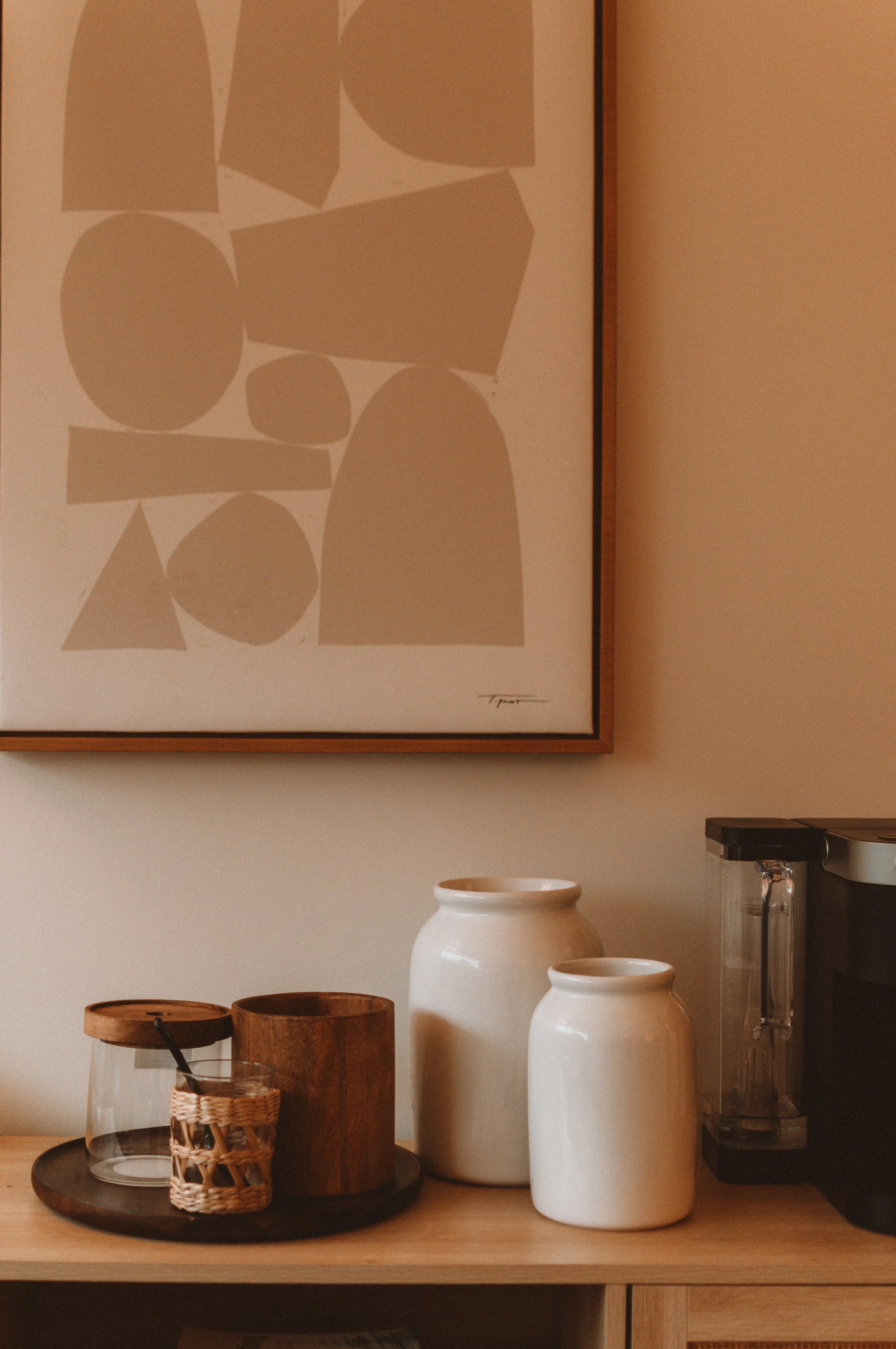 A wooden surface with two white vases, a glass jar with a wooden lid, a small woven basket, and a wooden container, positioned under an abstract artwork with geometric shapes, in a cozy interior setting.