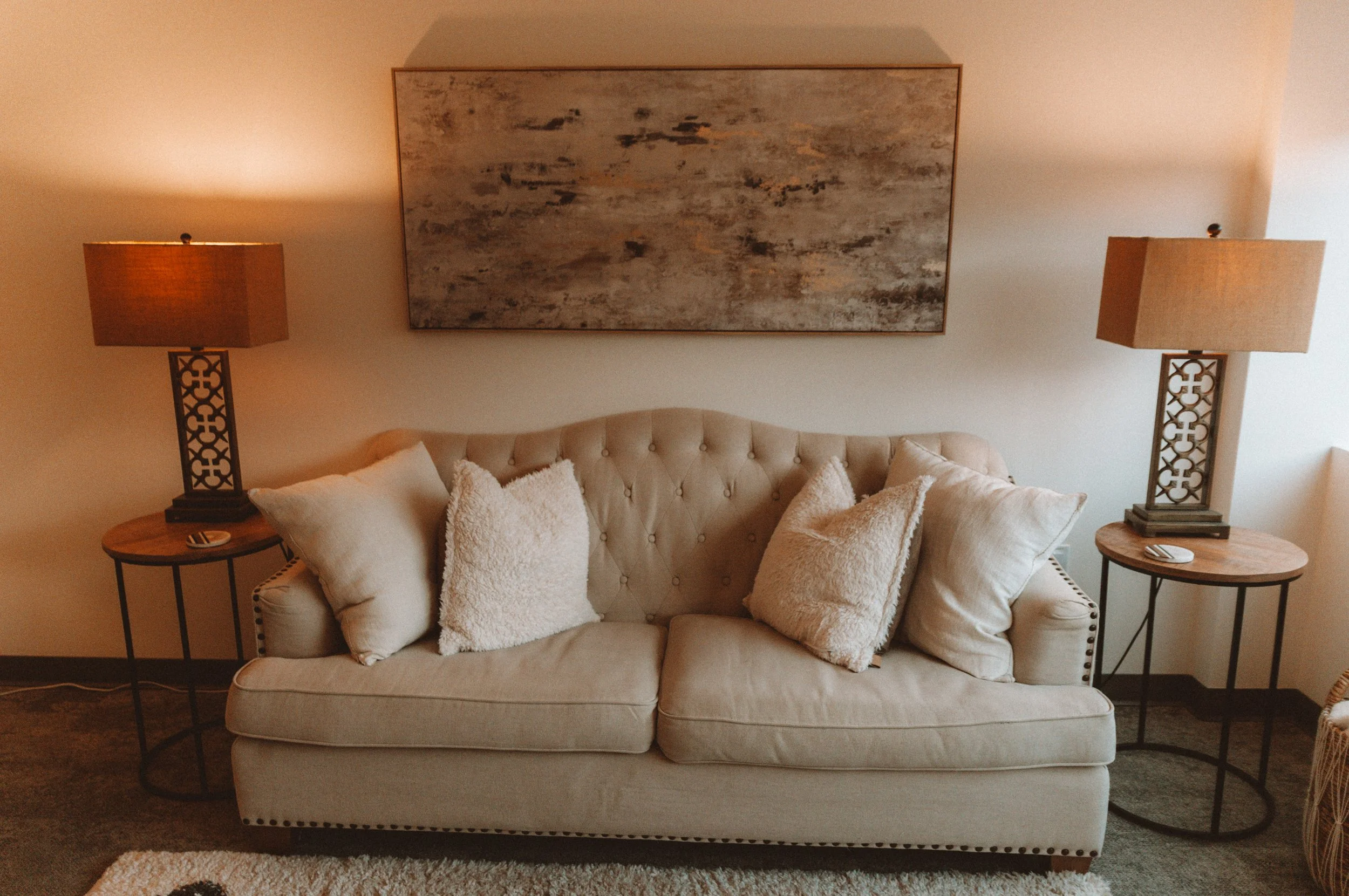 Living room with a beige tufted couch, six cream pillows, two wooden side tables with lamps, a large abstract painting on the wall, and a beige rug.