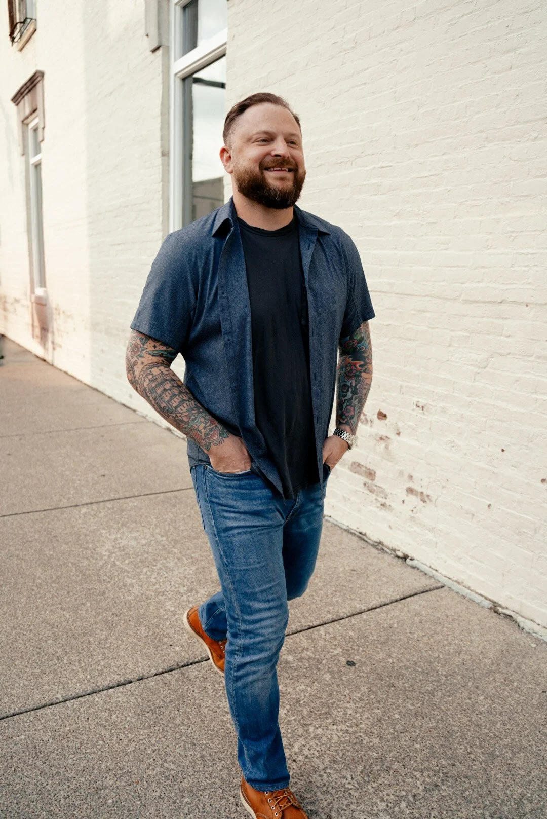 A man with tattoos on both arms walking outdoors, smiling, wearing a short-sleeved denim shirt over a black t-shirt, blue jeans, and orange shoes, against a white brick wall.