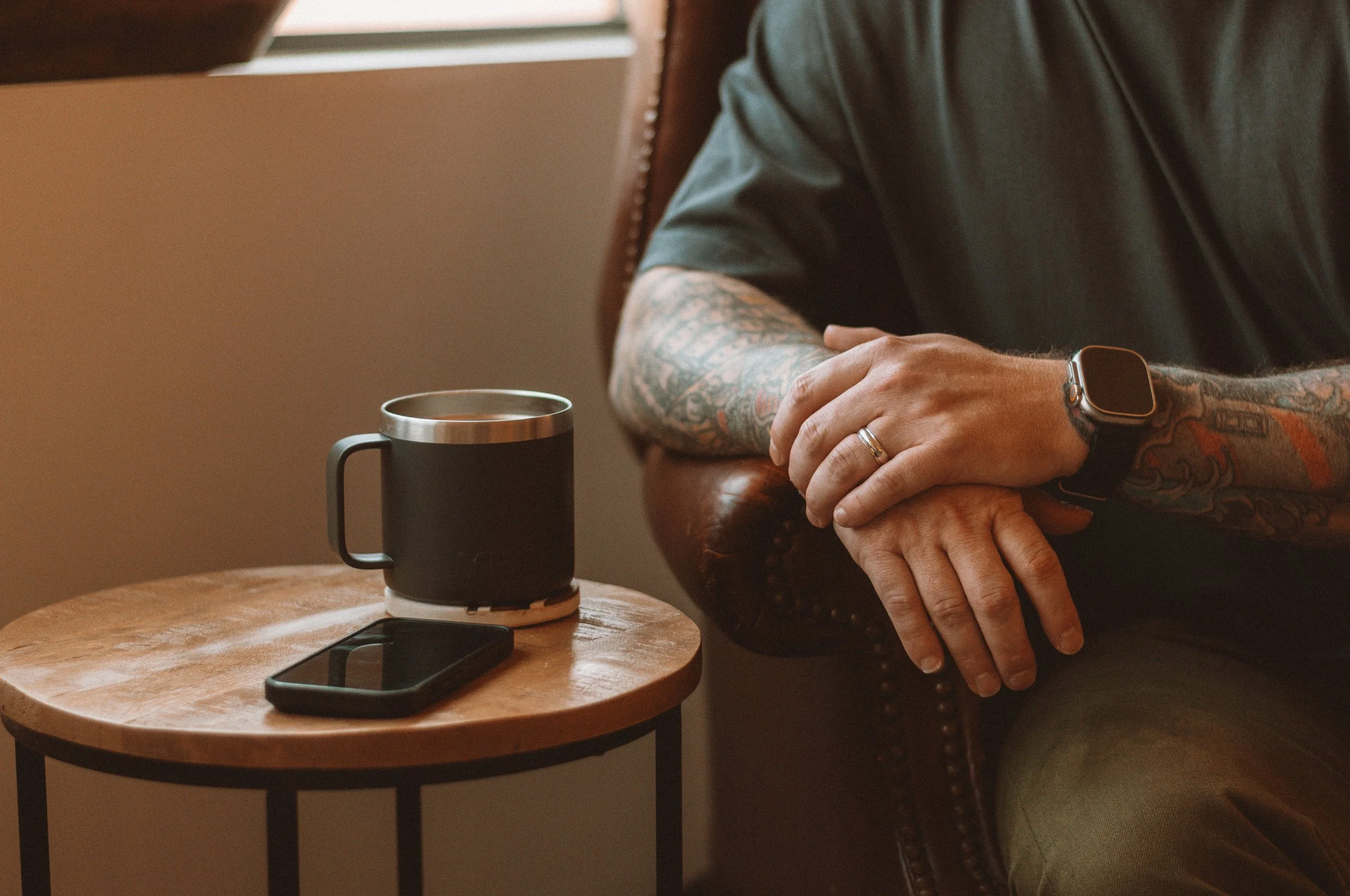 Person with tattoos on arms sitting on a brown leather chair near a round wooden side table with a black cup, a smartphone, and a smartwatch.